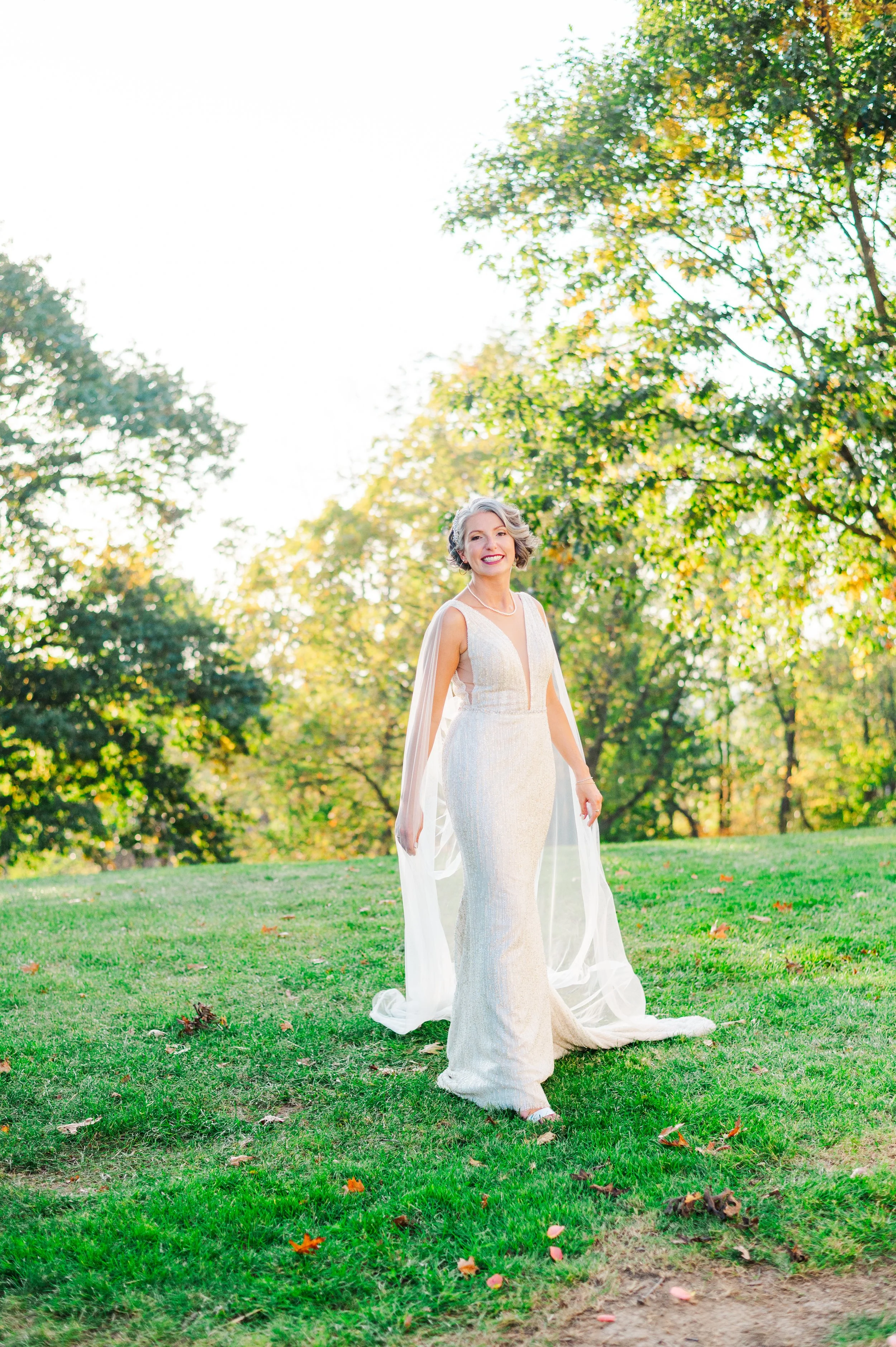 A woman in a white wedding gown standing on grass in a park with trees and sunlight in the background.