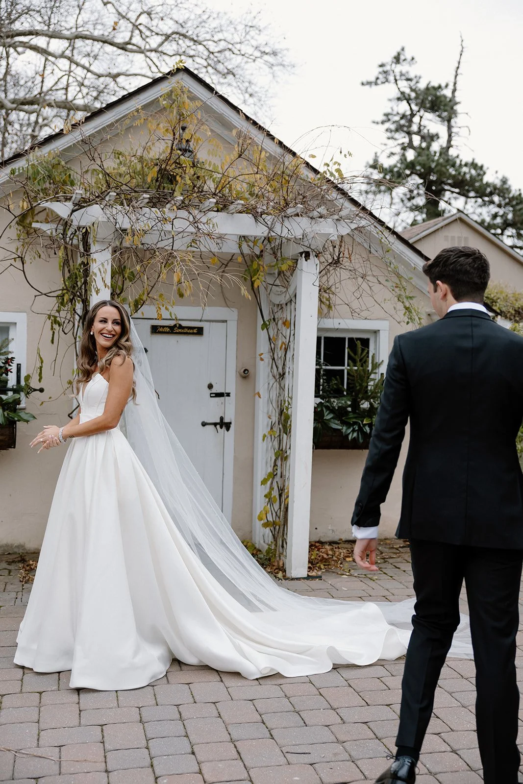 A bride in a white wedding gown and veil smiling at a groom in a black tuxedo outside a house with vines growing on the porch. strapless satin ballgown wona concept bridal dress
