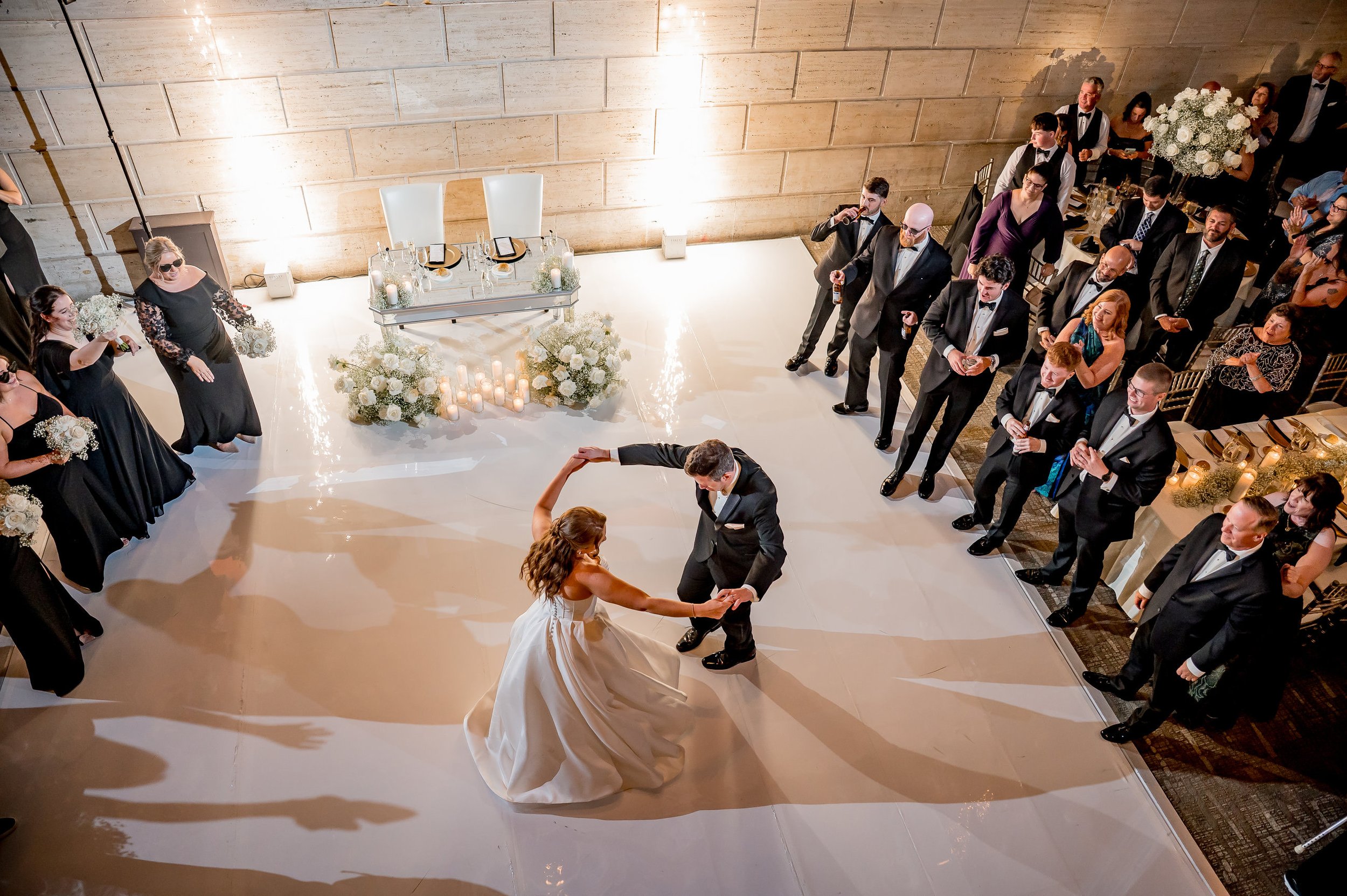 A bride and groom dance on the floor while guests in formal attire watch during a wedding reception.