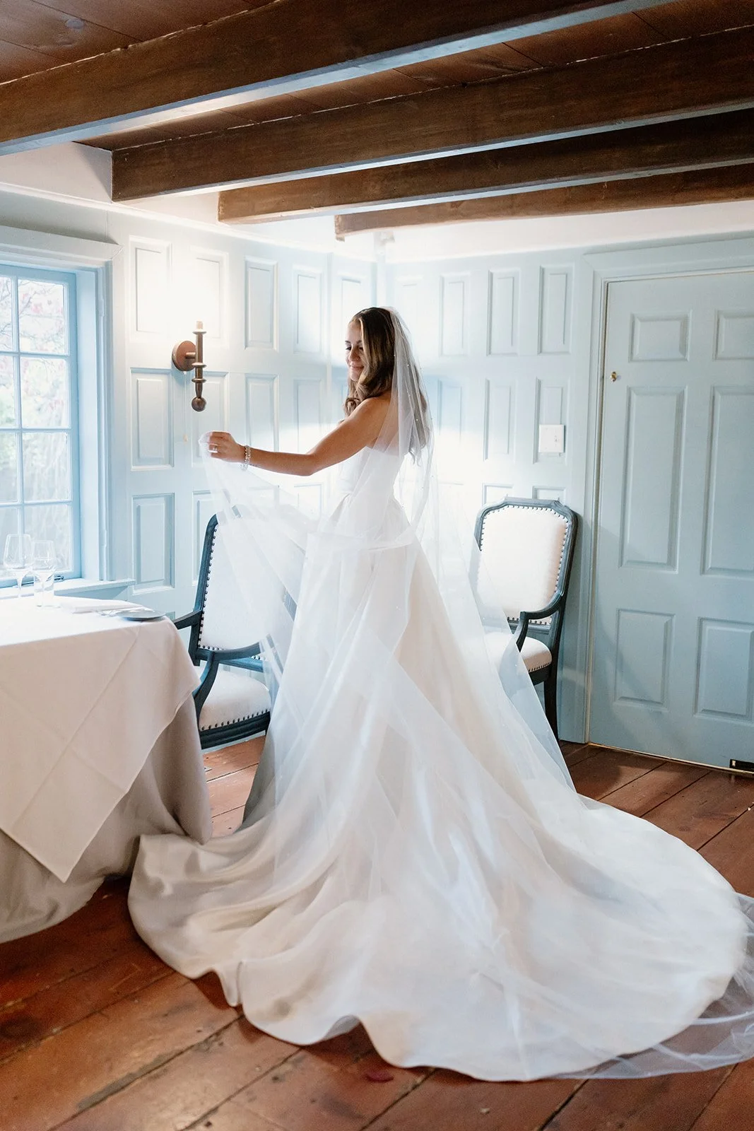 A bride in a white wedding gown adjusting her veil in a bright room with wooden beams on the ceiling and white paneled walls. strapless satin ballgown wona concept bridal dress