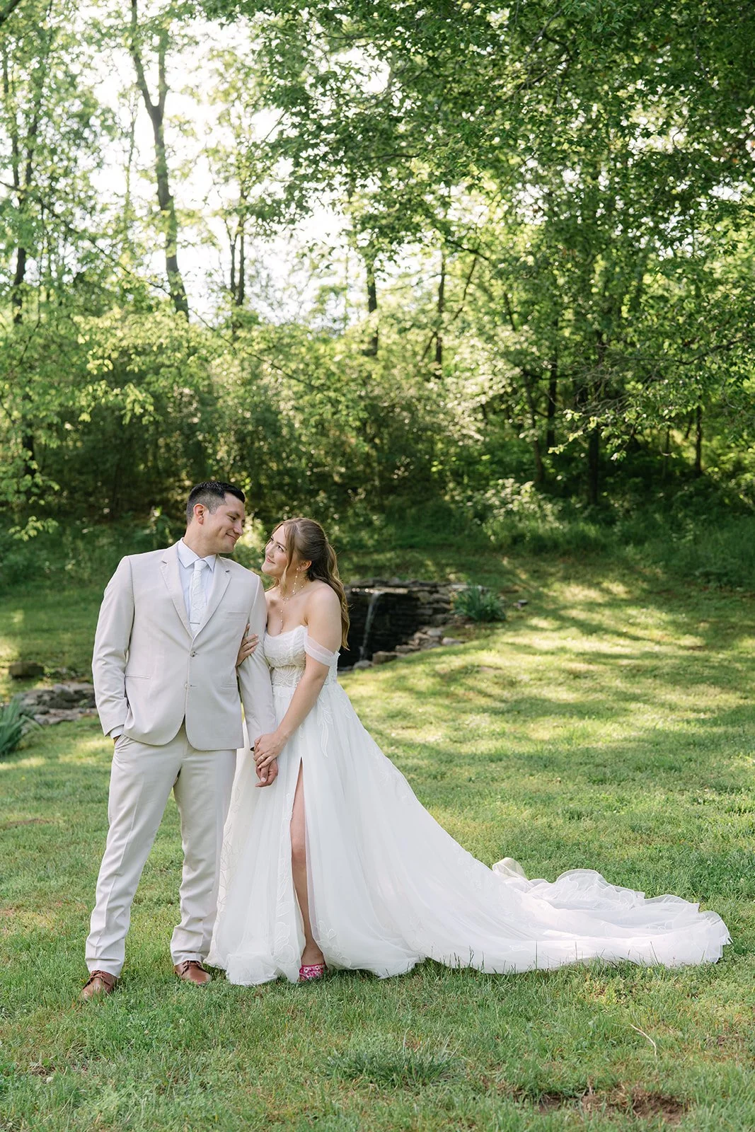 A bride and groom standing together outdoors in a lush green park with a small waterfall in the background. Tara Lauren De Mille