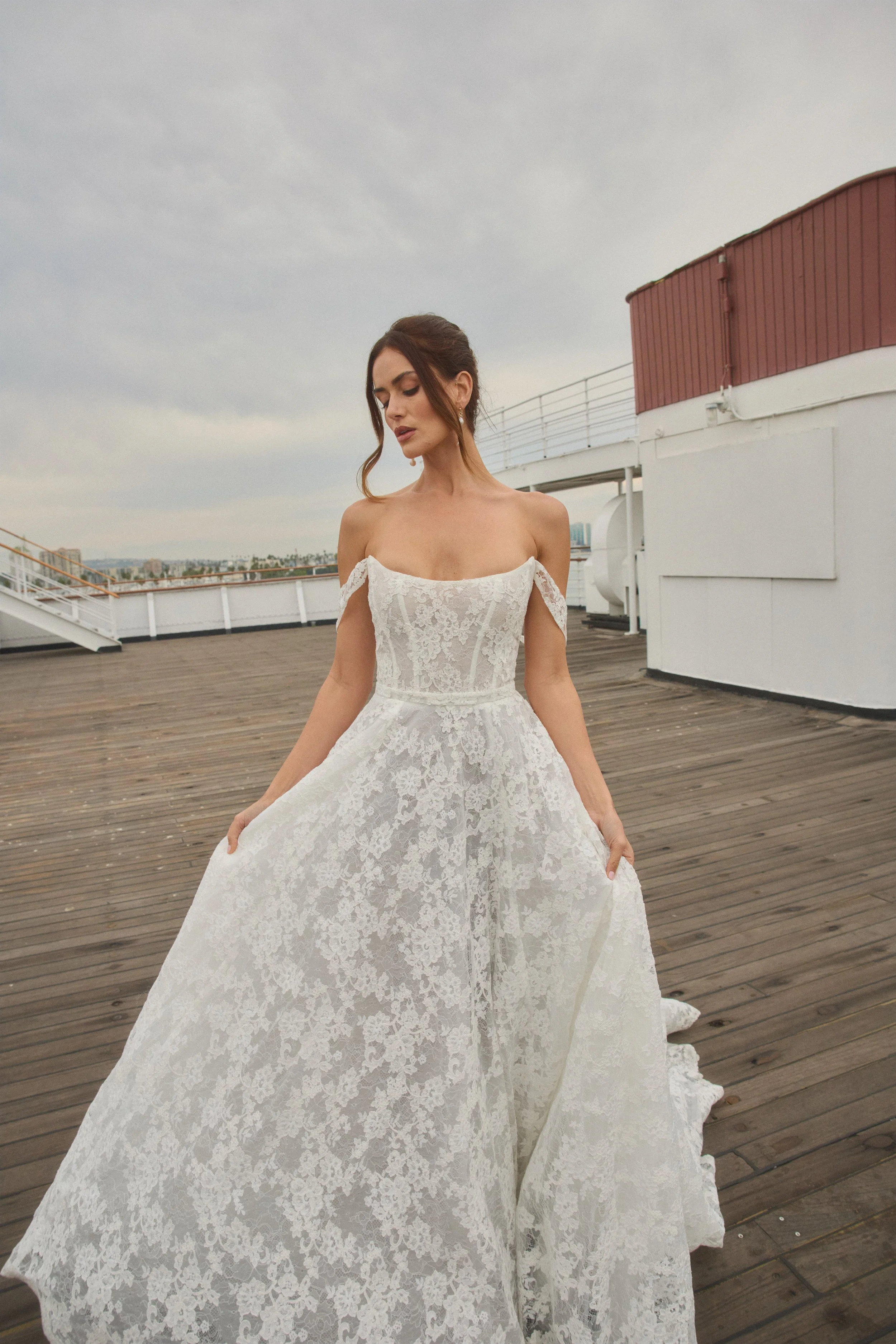 A woman in a white lace off-the-shoulder dress on a rooftop with a cloudy sky. tara lauren
