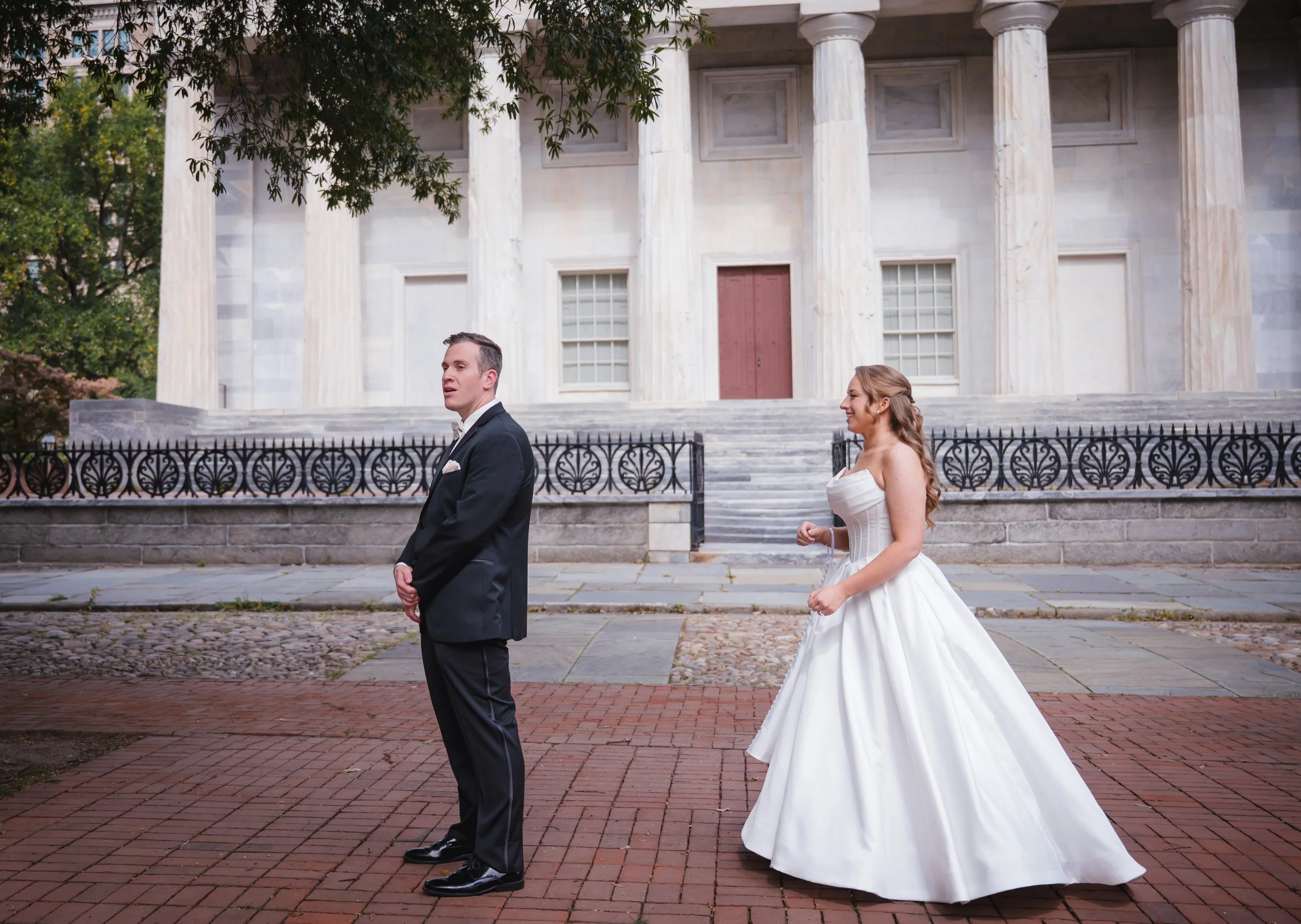 A bride and groom are standing outdoors in front of a historic building with columns, stairs, and iron railing. The bride is in a white wedding gown and the groom in a black tuxedo.