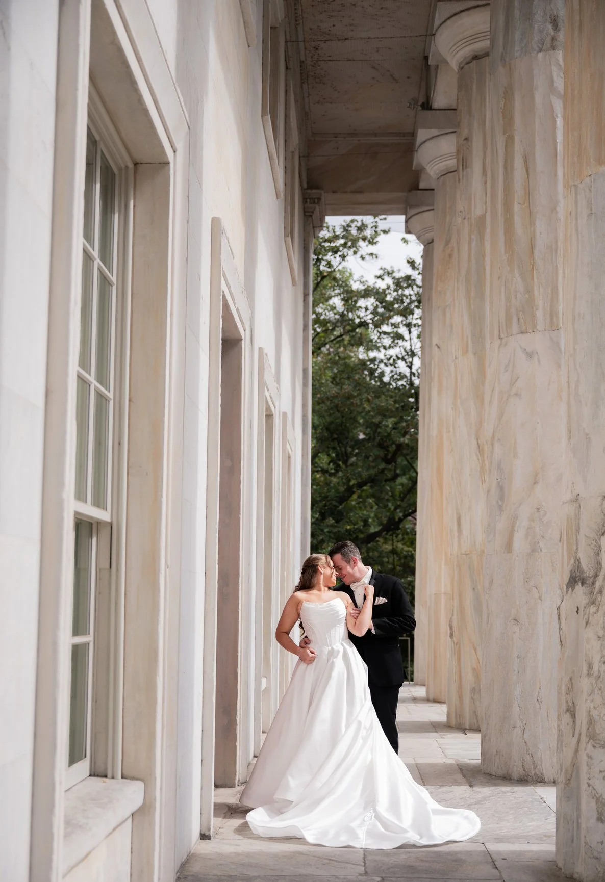 A bride and groom dancing closely under a marble portico with large columns and windows, with green trees in the background.