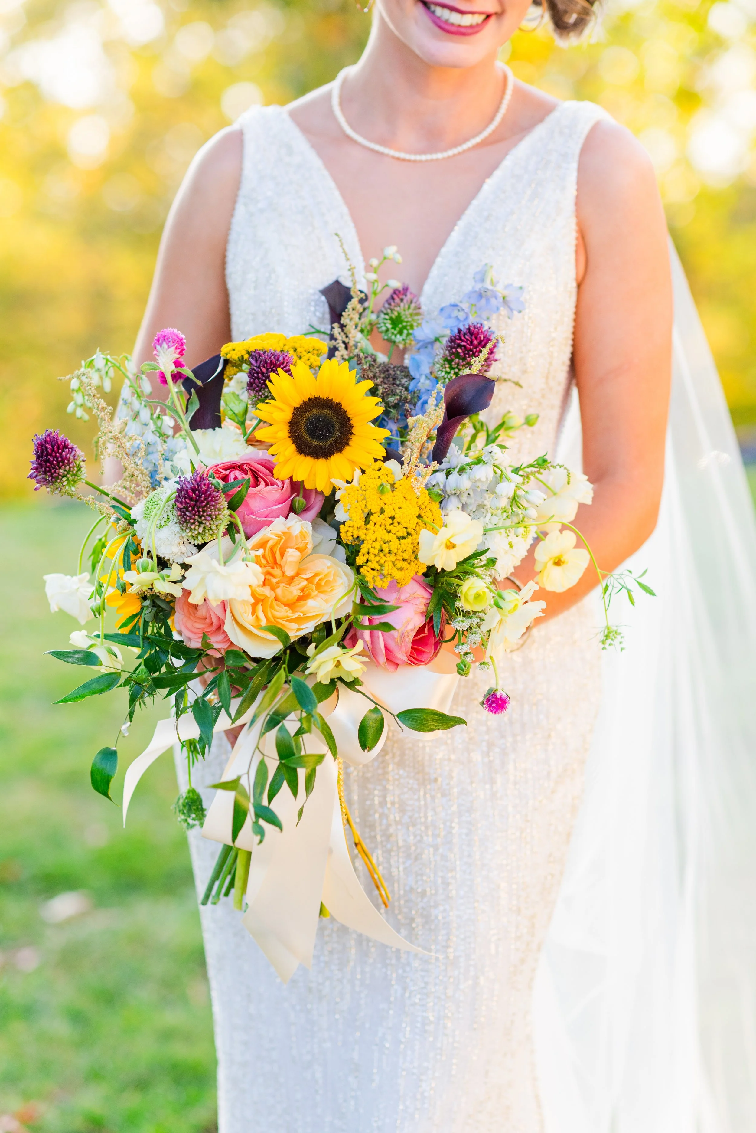 Bride holding a colorful bouquet of flowers outdoors, wearing a white dress and pearl necklace, with sunlight and blurred greenery in the background.