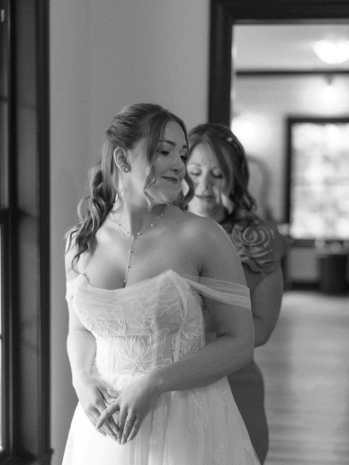 A woman in a wedding dress with a veil, holding her hands in front of her, smiling and looking down. Another woman behind her appears to be adjusting the dress. Tara Lauren De Mille