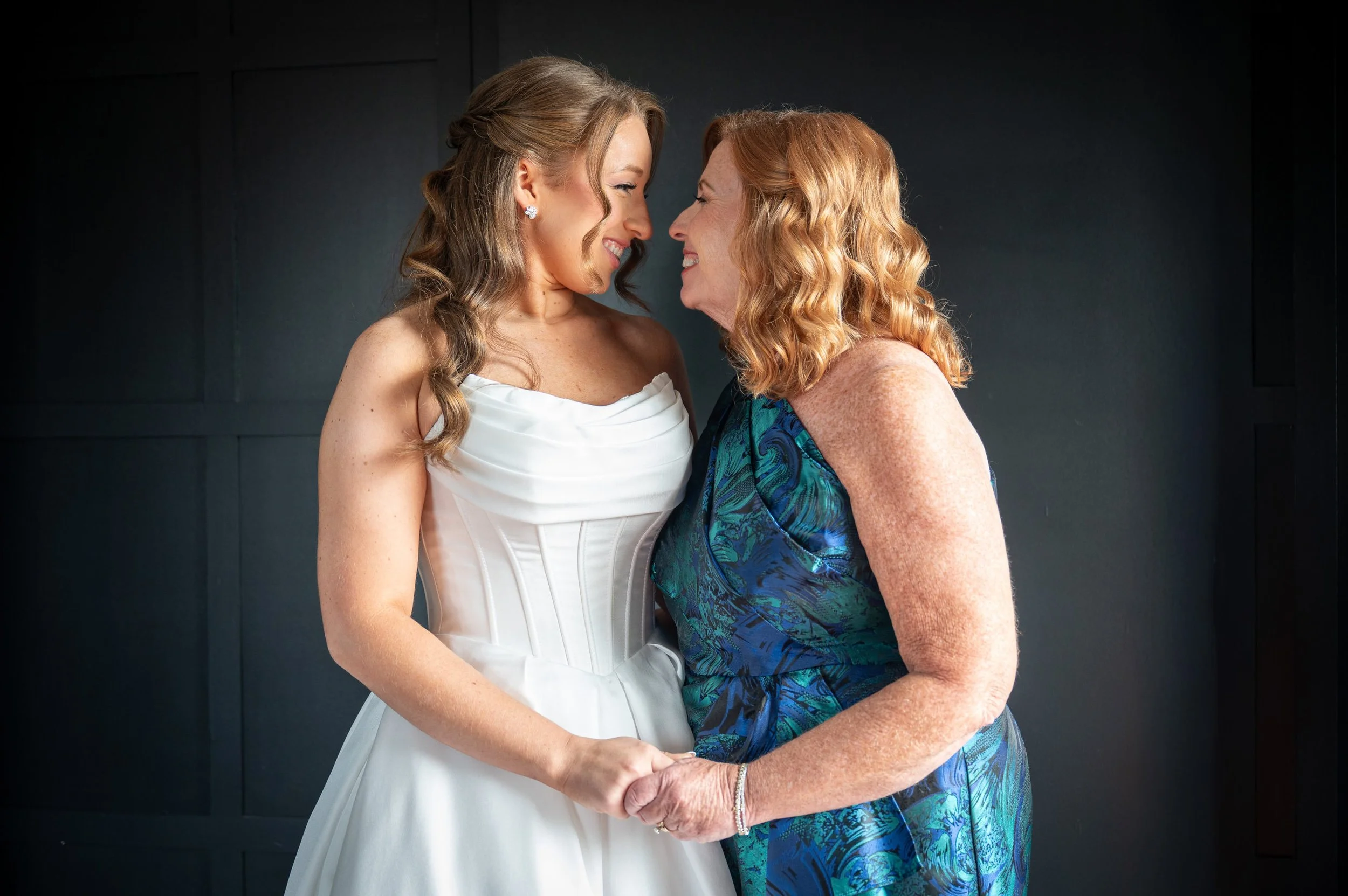 A woman in a white wedding dress and a woman in a blue patterned dress smiling and holding hands against a black background.