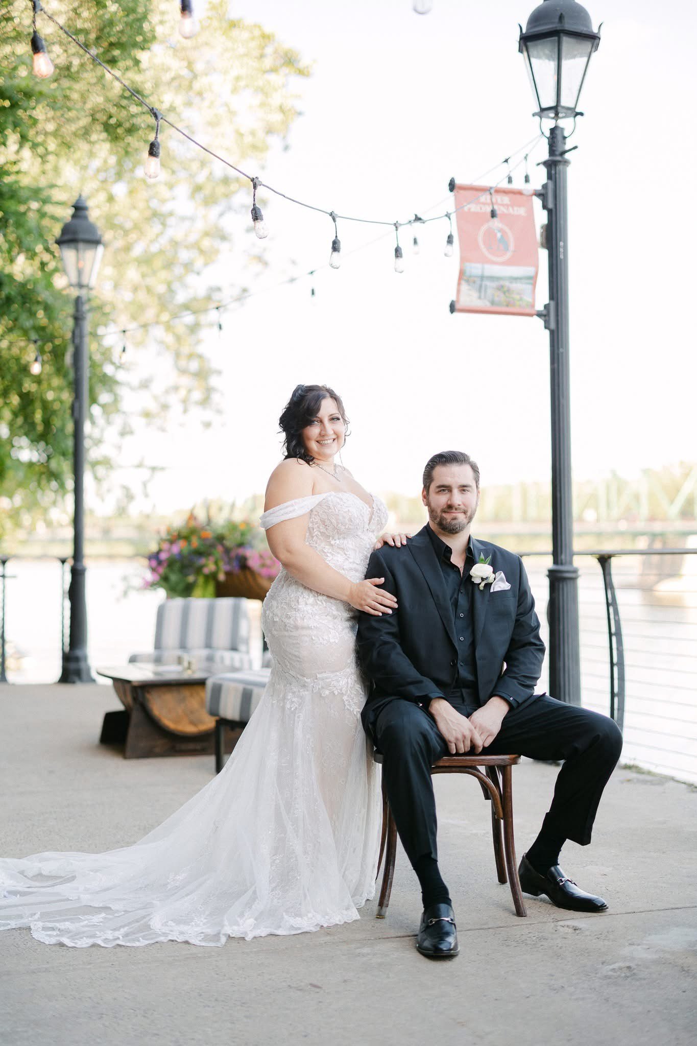 A bride and groom pose outdoors on a riverfront walkway with trees, string lights, and a decorative lamp post.