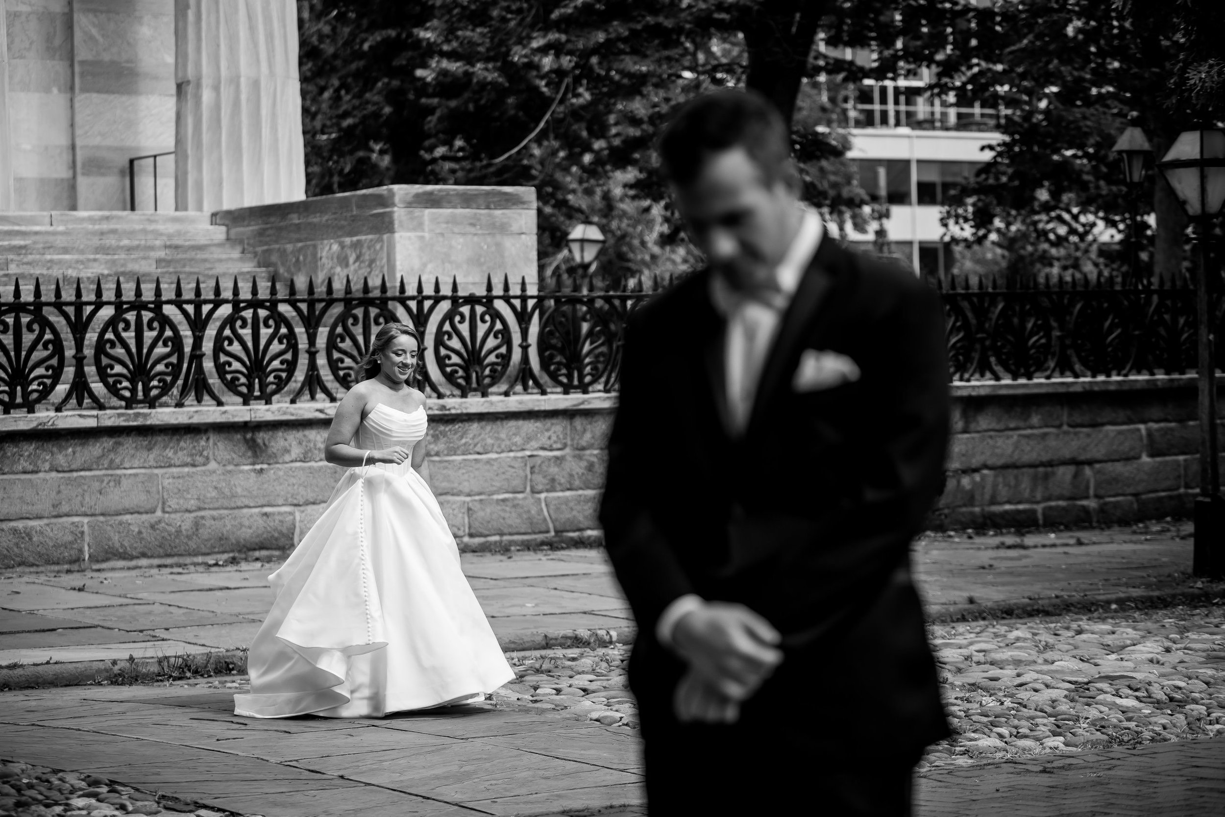 A bride in a white wedding gown smiling at a groom in a dark suit, standing outdoors near a stone fence and trees in black and white.
