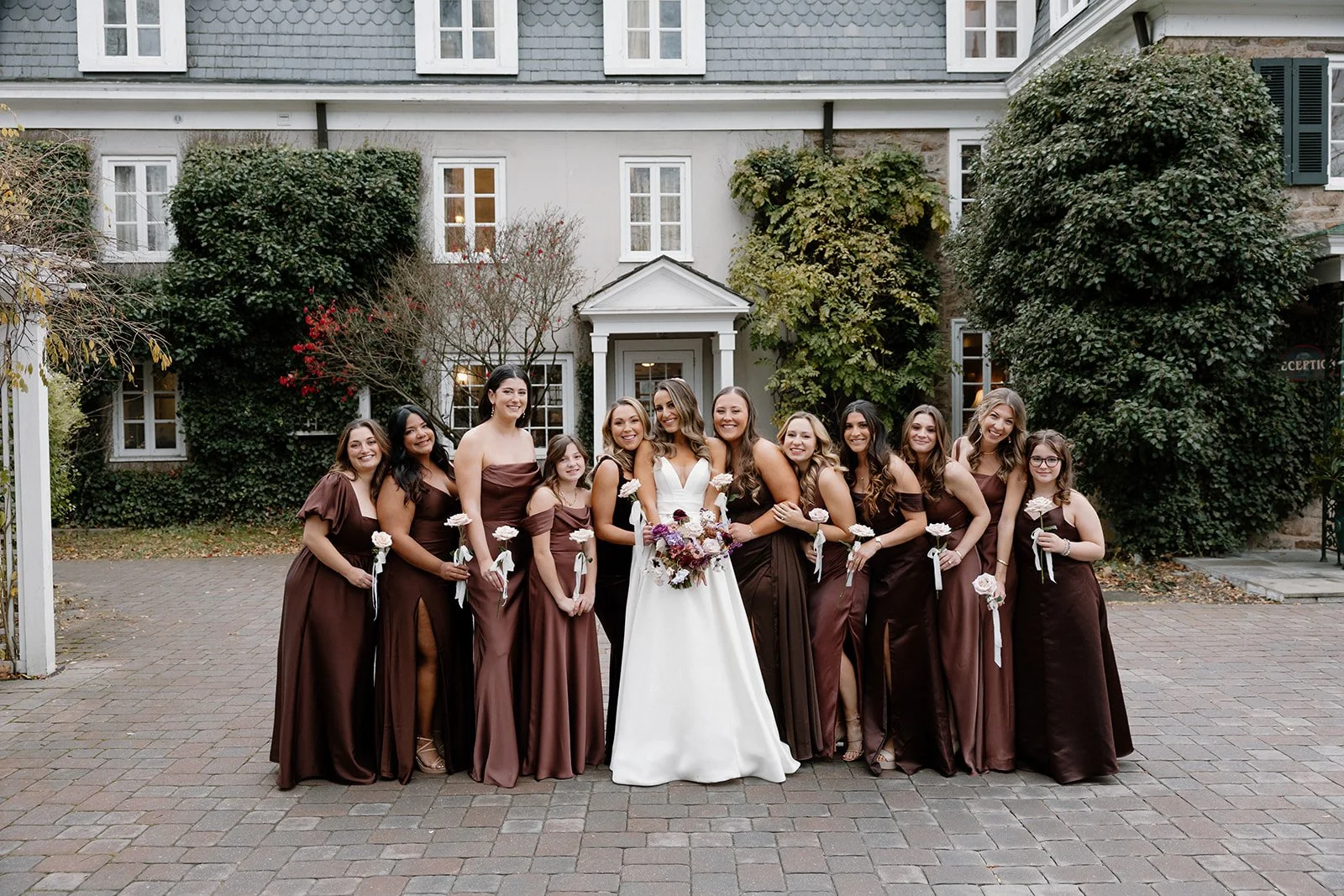 A bride in a white wedding dress and her bridesmaids in matching brown dresses stand together outdoors in front of a house with ivy-covered walls. strapless satin ballgown wona concept bridal dress