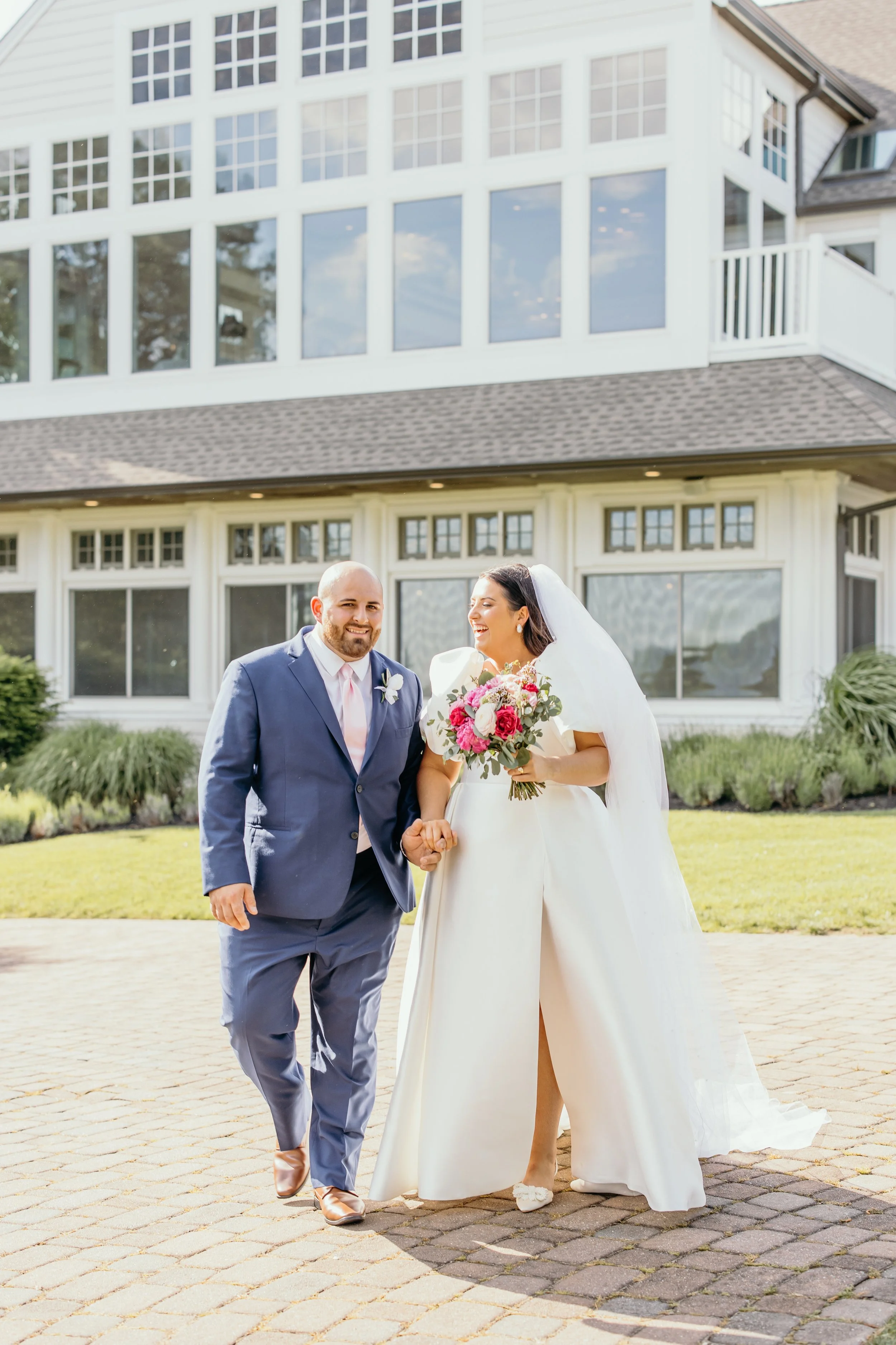 A bride and groom walking hand-in-hand outside a large, white, multi-story house with big windows. The bride is wearing a white dress with a slit, holding a bouquet of pink and white flowers, and smiling. The groom is wearing a navy suit with a pink tie, smiling as well.