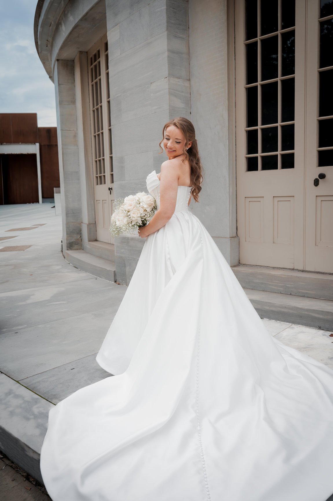 A bride in a white wedding gown holding a bouquet of white flowers, standing outside a building with large windows and stone walls.