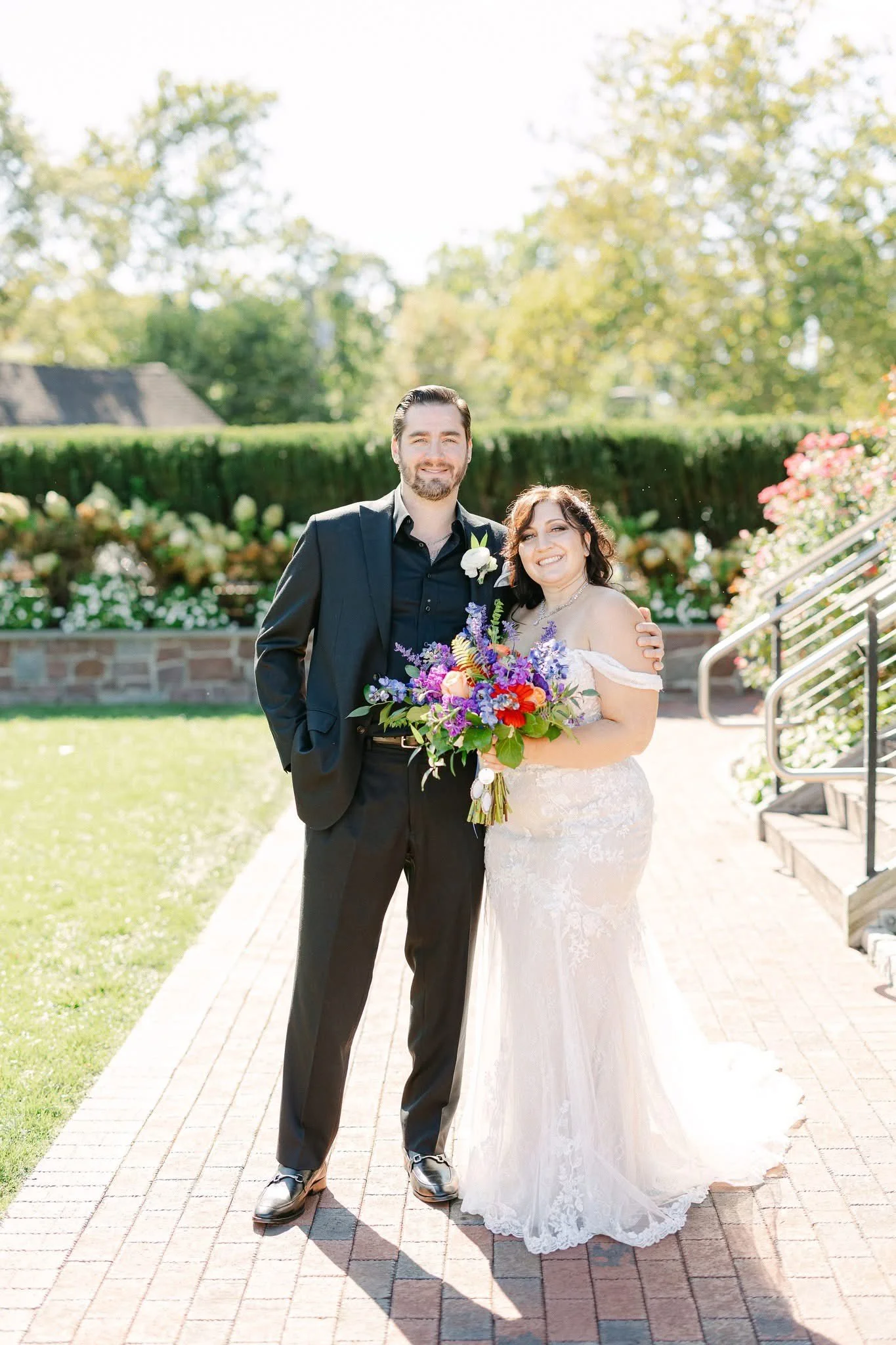 A bride and groom standing outdoors on a sunny day. The bride is holding a colorful bouquet of flowers and is dressed in a white lace wedding gown. The groom is in a dark suit with a boutonniere. They are smiling and embracing. In the background, the