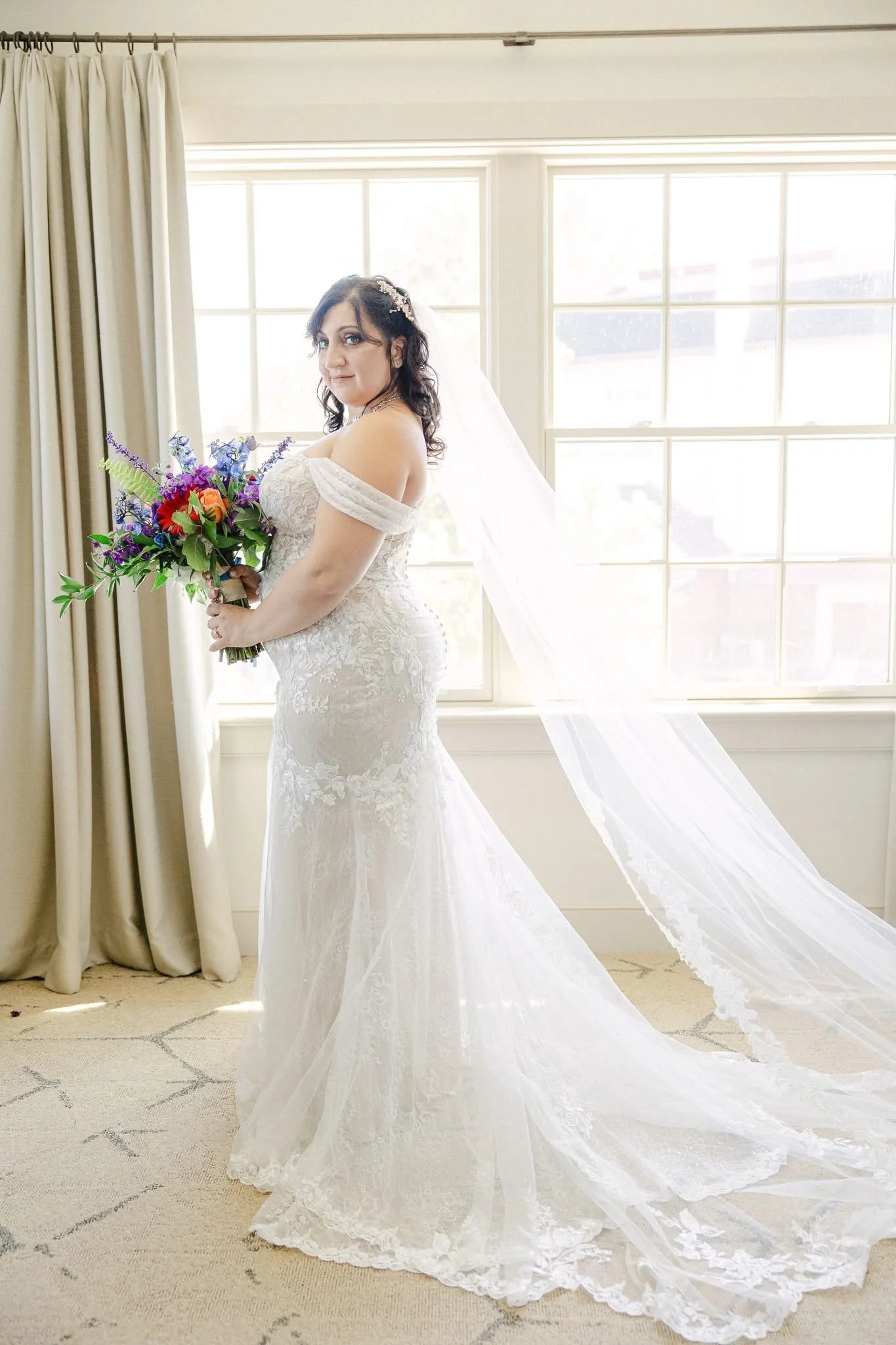 Bride in a wedding dress holding a colorful bouquet, standing in front of a bright window with curtains.