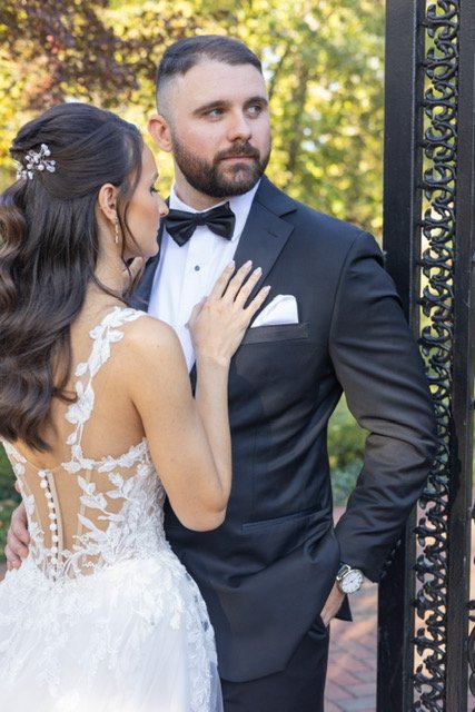 A bride in a white wedding gown touching a groom in a black tuxedo, standing outdoors near a black iron gate with trees in the background.
