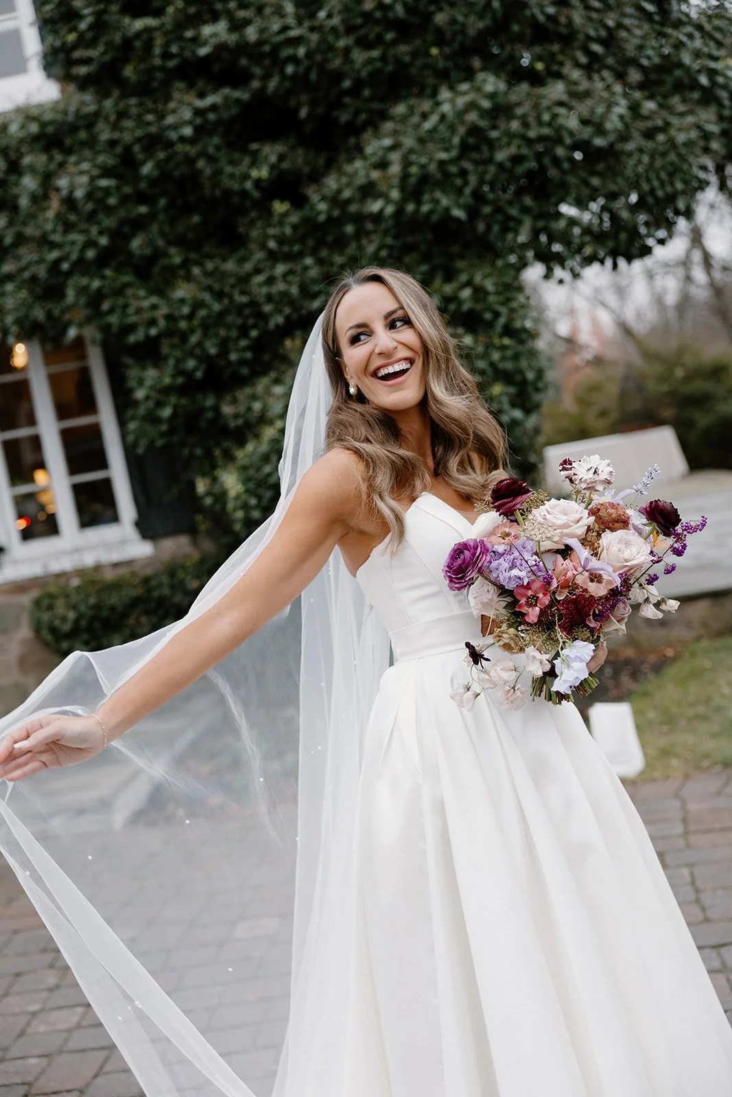 A smiling bride in a white wedding dress holding a bouquet of purple, pink, and white flowers outdoors. strapless satin ballgown wona concept bridal dress