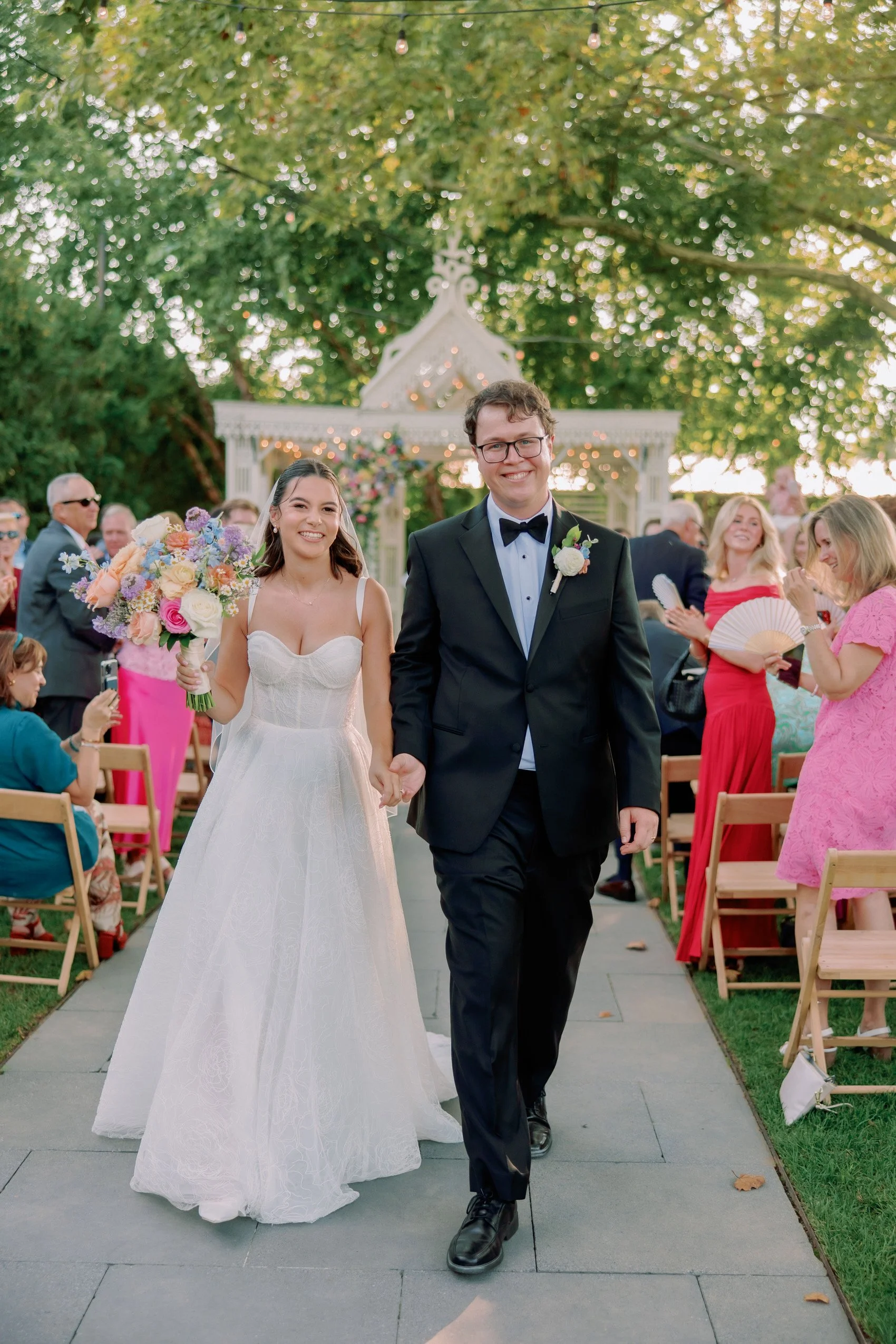 A bride and groom walking hand in hand down an outdoor wedding aisle, smiling, with guests clapping and taking photos around them, and a decorative gazebo in the background with trees and string lights. Tara Lauren Kismet