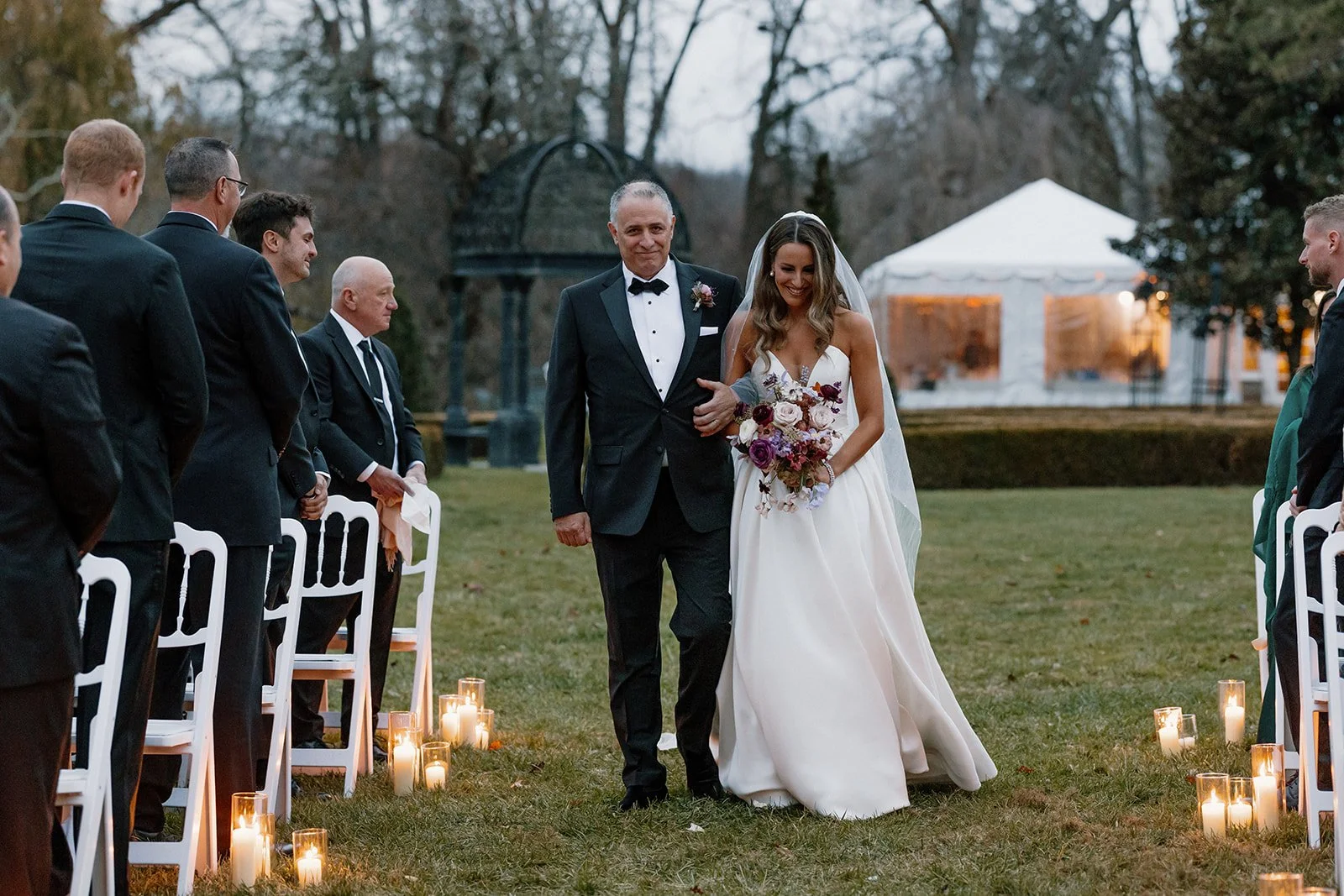 Bride and her father walking down the outdoor wedding aisle in the evening, surrounded by guests dressed in formal attire, with candles lining the aisle and a large white tent in the background. strapless satin ballgown wona concept bridal dress