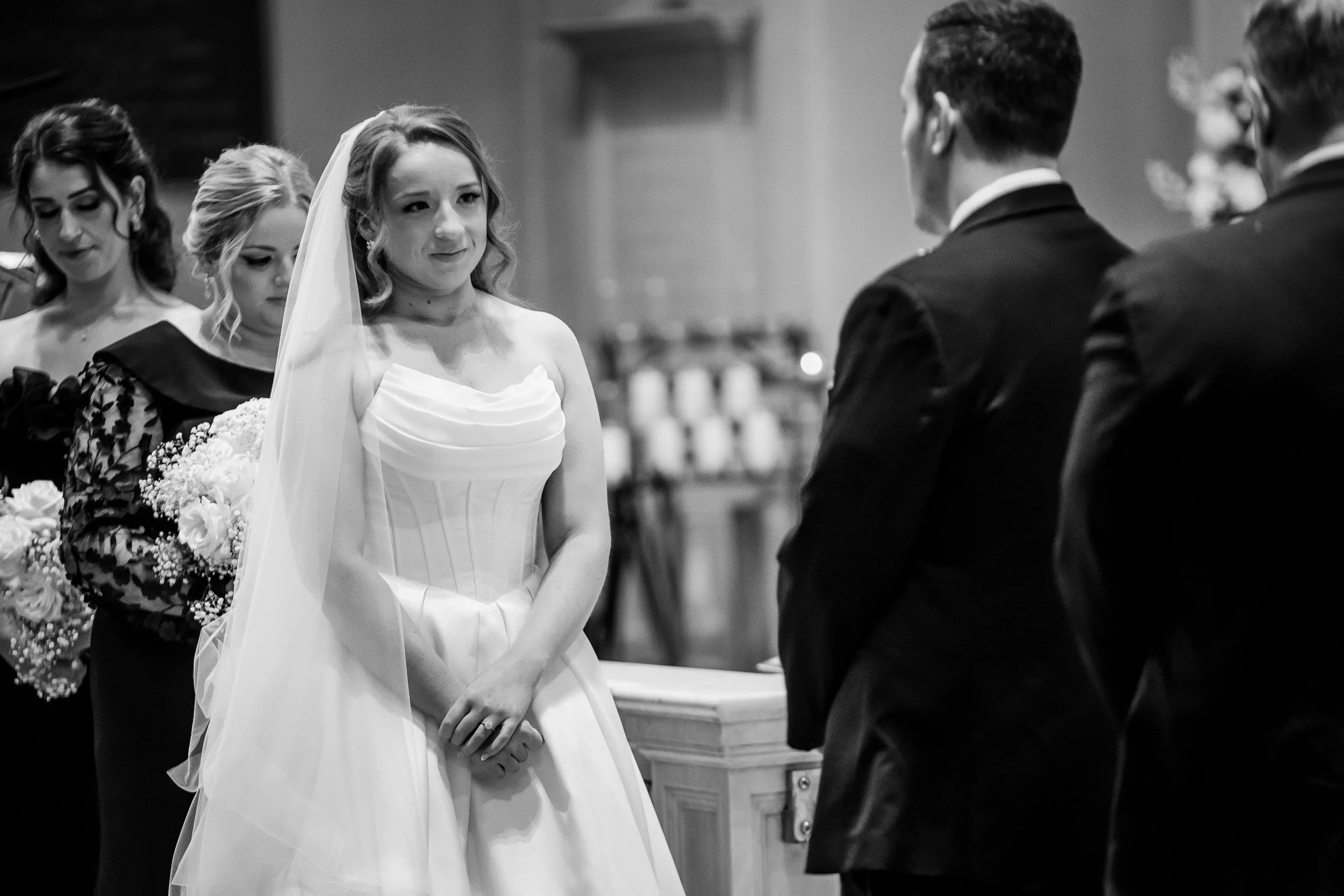 A bride standing before her groom during a wedding ceremony, with bridesmaids behind her and groomsmen in front, in a church setting.