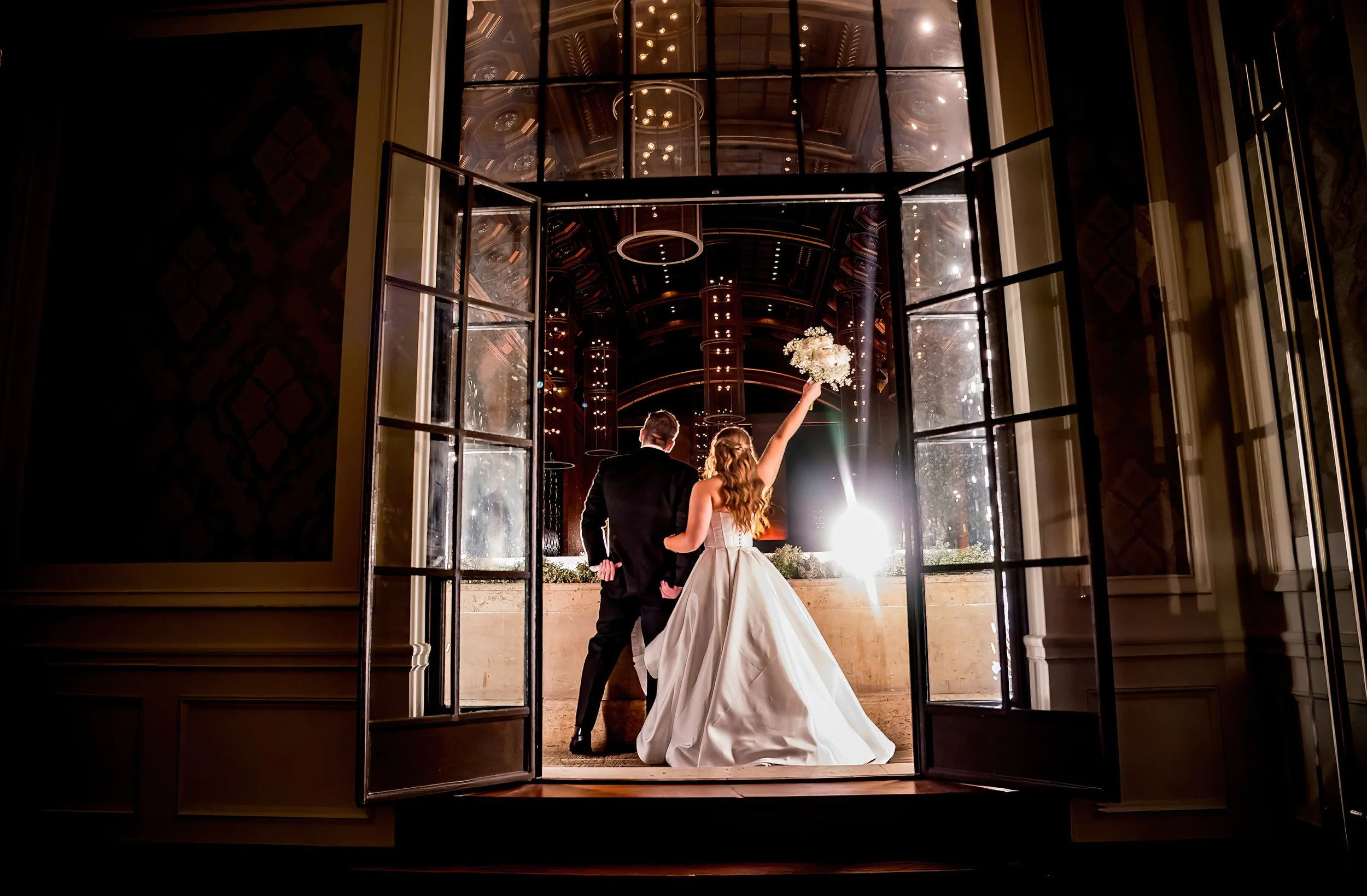 Bride and groom standing at the window of an elegant venue, with the bride holding a bouquet of flowers and raising her arm in celebration.