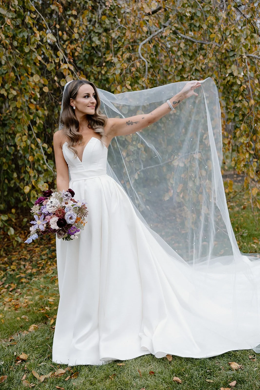 A bride in a white wedding dress holding a bouquet of flowers outdoors with trees in the background. strapless satin ballgown wona concept bridal dress