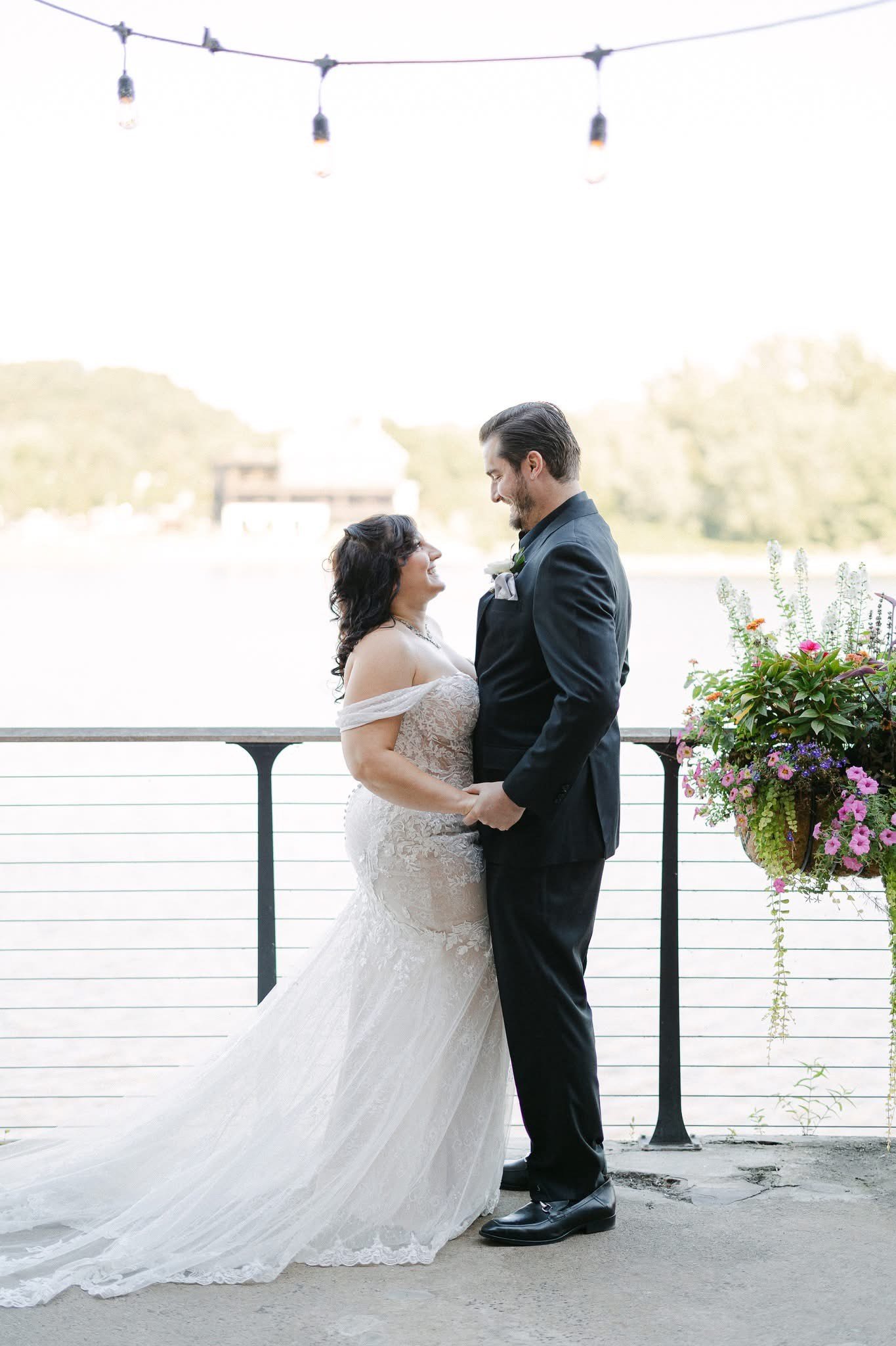 A bride and groom are holding hands and smiling at each other during their wedding by a waterfront with string lights overhead and flower arrangements nearby.