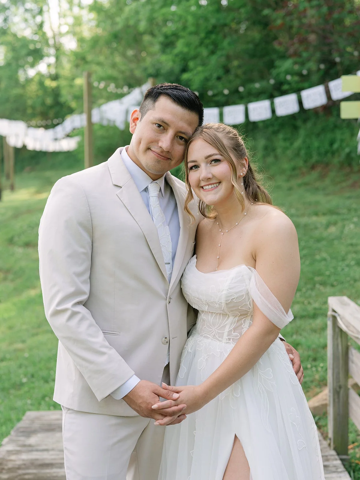 A newlywed couple in white wedding attire stands outdoors on a wooden bridge, holding hands and smiling at the camera, with green trees and hanging decorations in the background. Tara Lauren De Mille