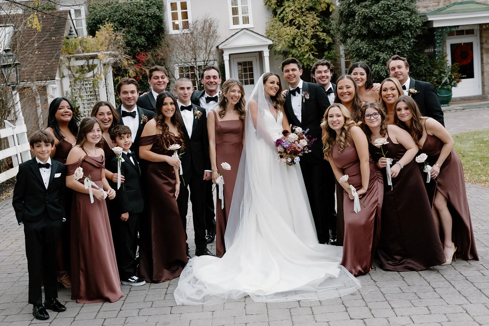 A large wedding party outdoors, featuring a bride and groom in the center, surrounded by bridesmaids and groomsmen, all smiling and dressed formally, with a house and greenery in the background. strapless satin ballgown wona concept bridal dress