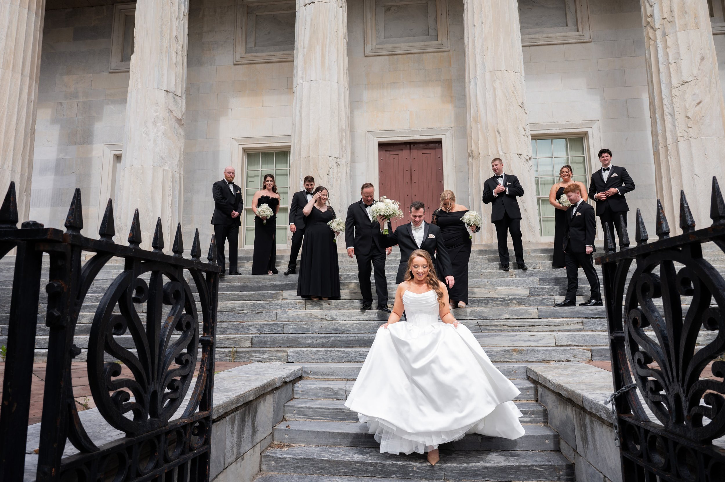 A bride in a white wedding dress with a full skirt and strapless bodice walking down stairs in front of a group of people dressed in black tuxedos and dresses, outside a large stone building with tall columns.