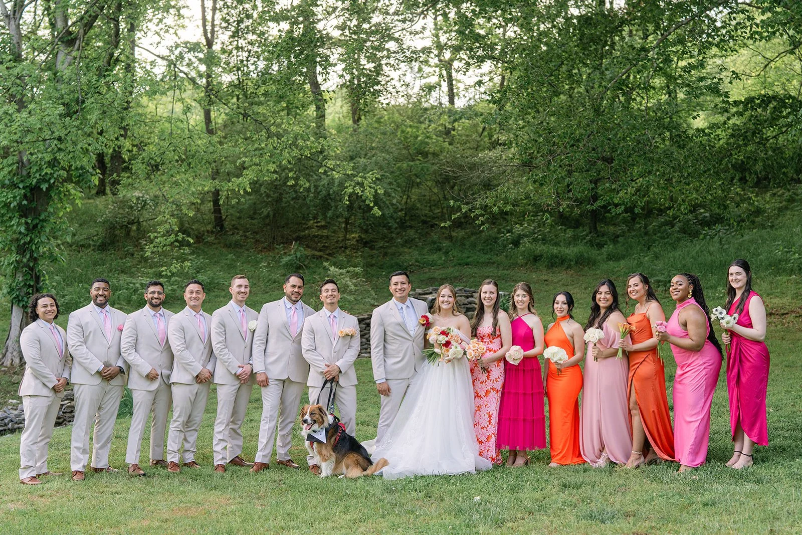 A wedding party outdoors on a grassy area with trees in the background, featuring the bride and groom, bridesmaids in pink and orange dresses, groomsmen in light-colored suits with pink ties, and a dog. Tara Lauren De Mille