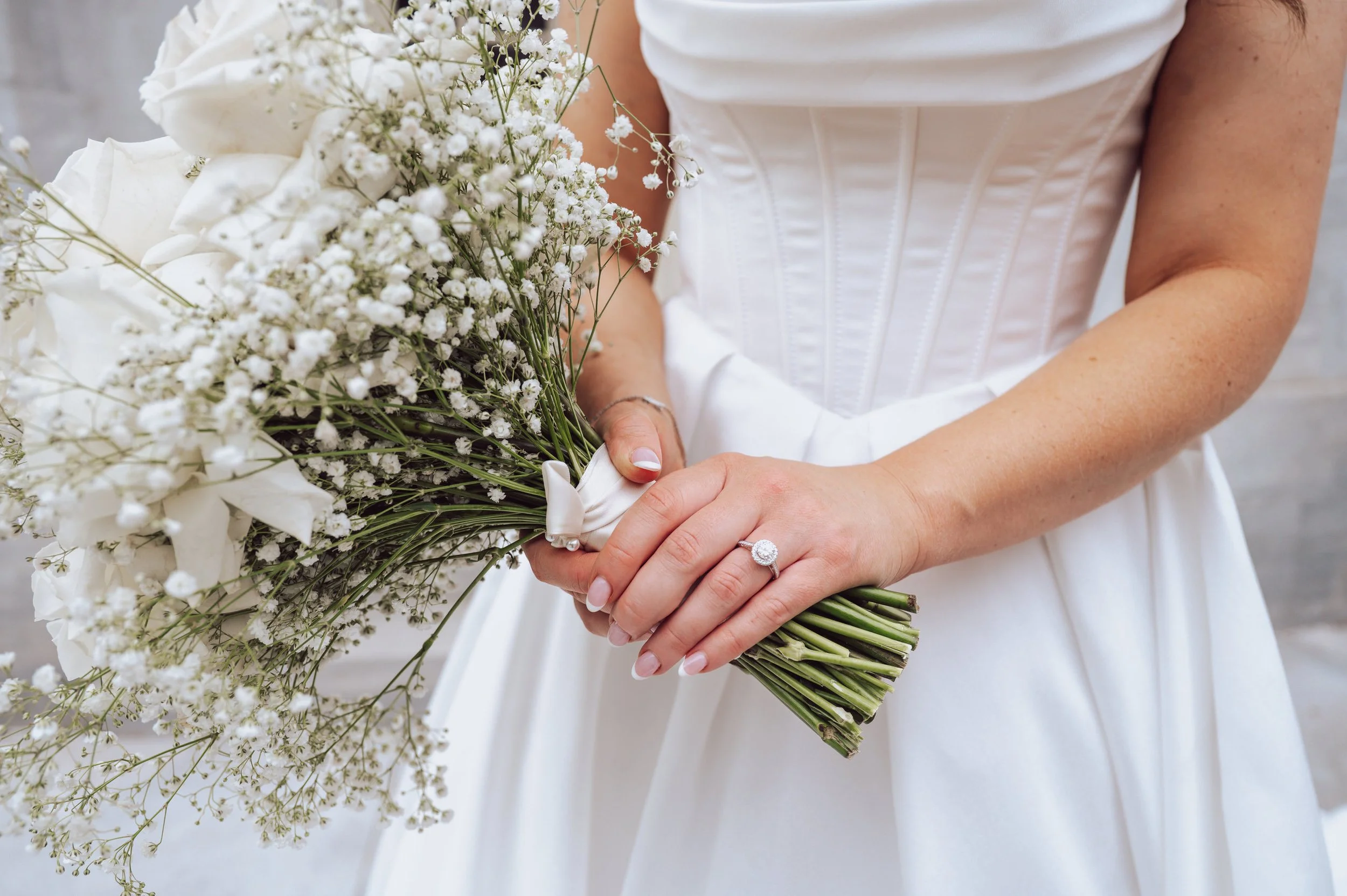 Close-up of a bride holding a bouquet of white flowers, including roses and baby's breath, with her hands clasped around the stems. She wears a diamond ring and a white wedding dress with a fitted bodice.