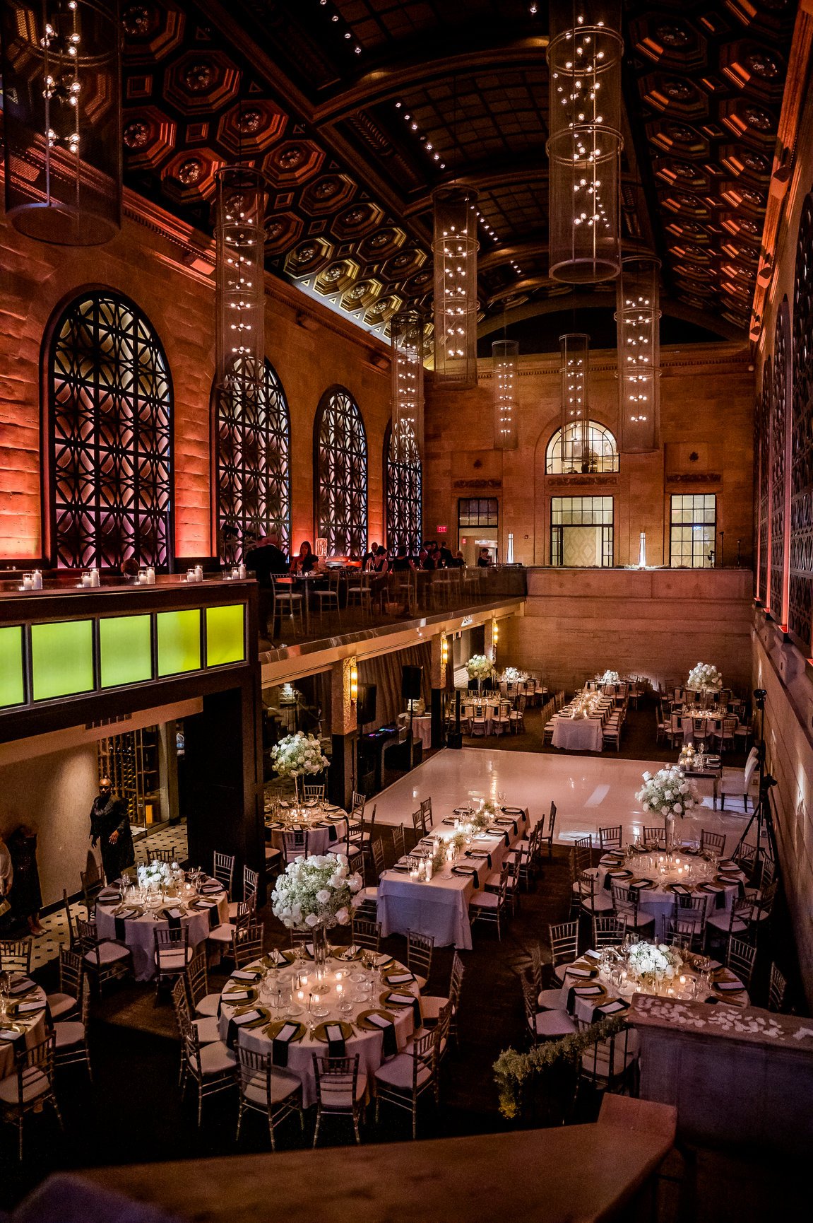 Elegant indoor event space decorated for a formal occasion, with round tables covered in pink tablecloths, floral centerpieces, and candles, arranged around a dance floor with a band or DJ setup, featuring warm lighting and tall windows with decorati