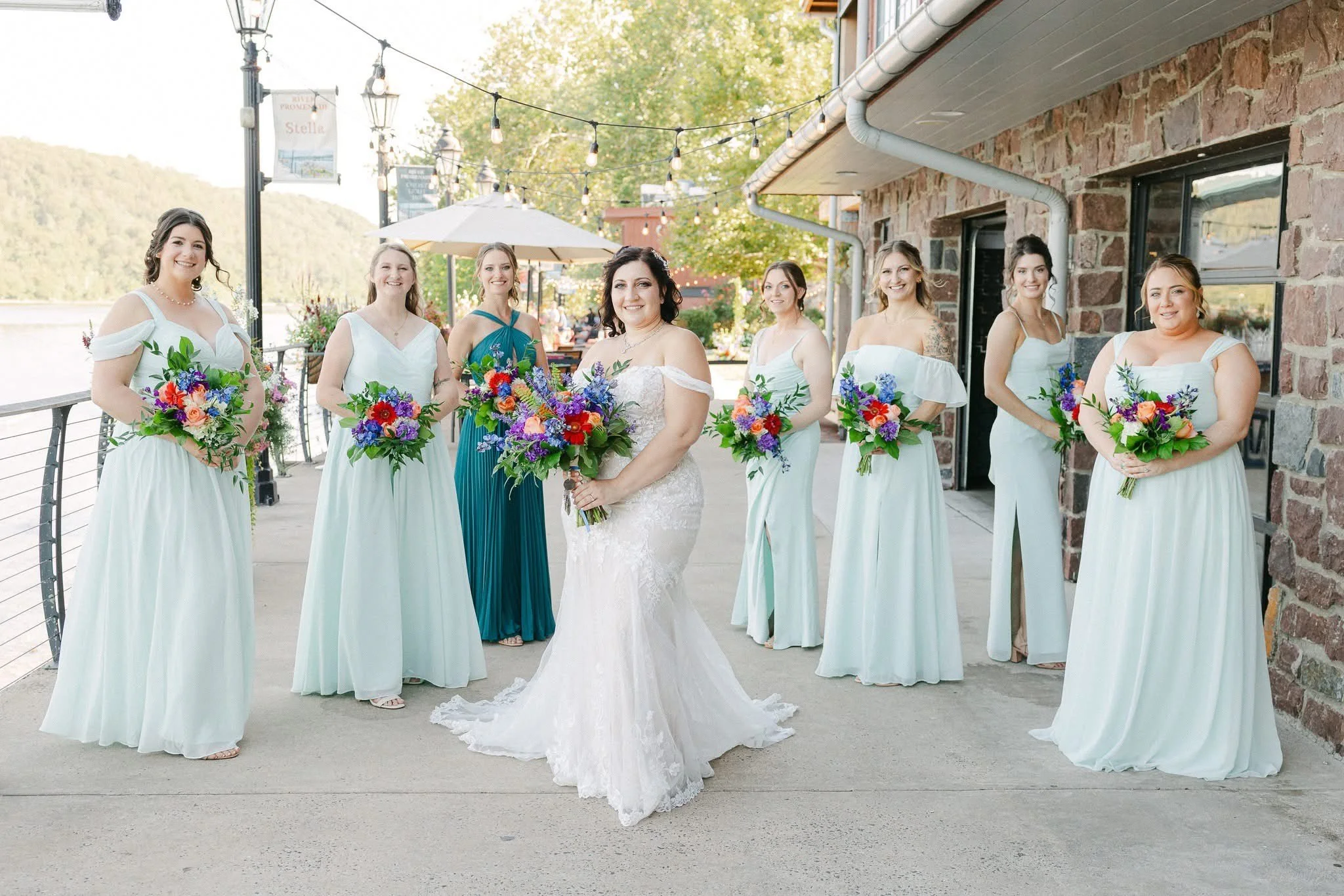 Bride in white wedding dress holding a colorful bouquet, surrounded by seven bridesmaids in light-colored dresses with colorful bouquets, standing outdoors near a stone building by a waterfront with string lights overhead.