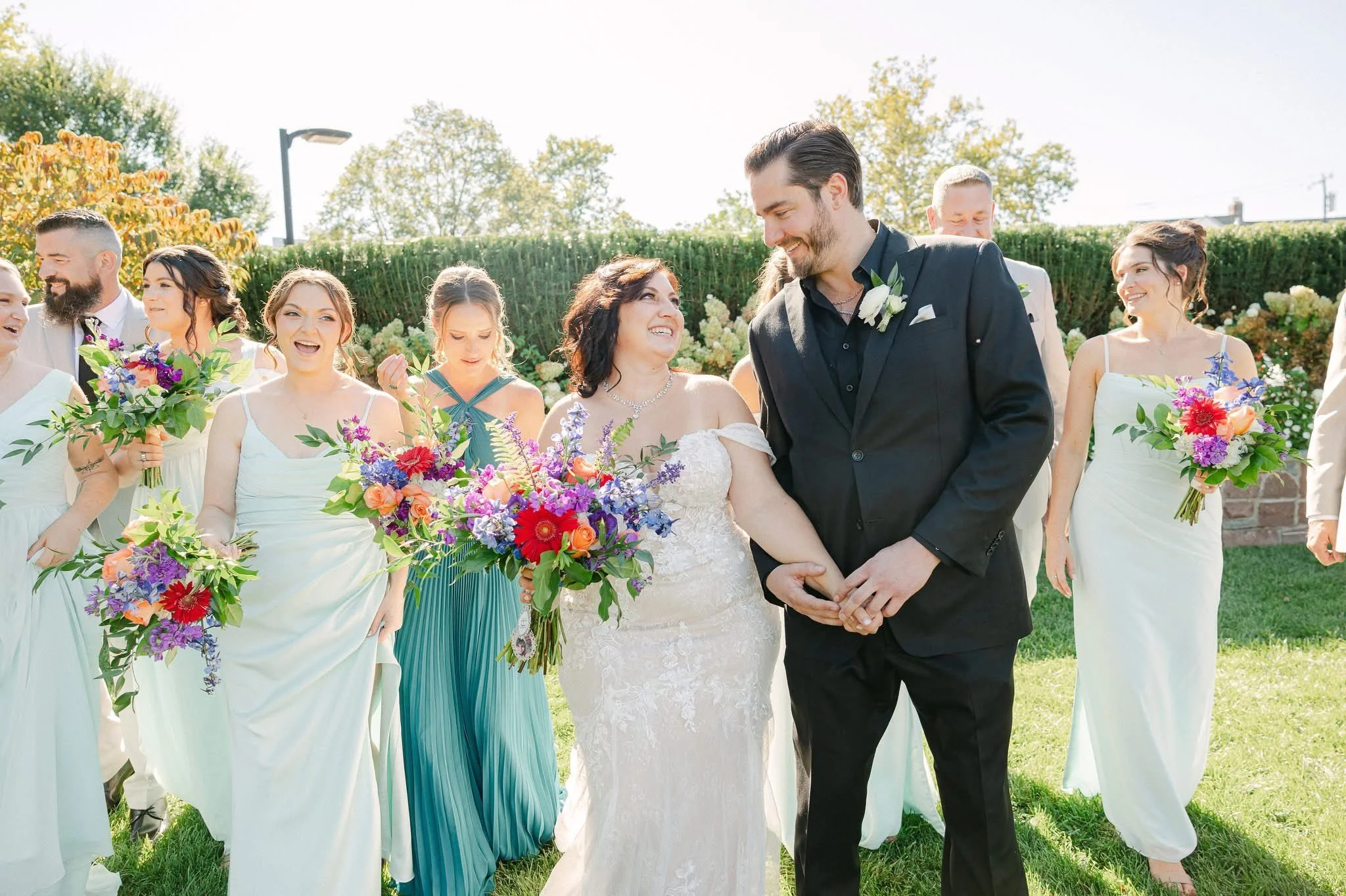 Bride and groom holding hands and smiling at each other, surrounded by bridal party with bouquets outdoors during wedding reception.