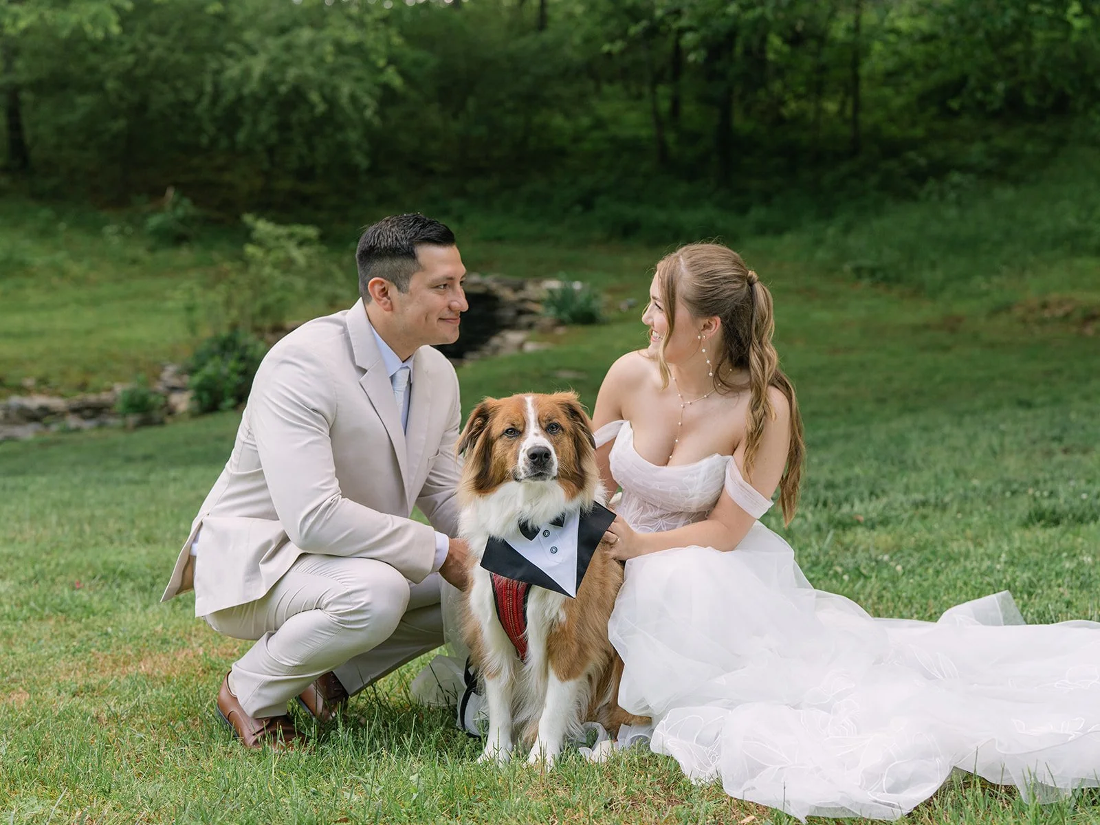 A couple in wedding attire with a dog in a park-like setting, smiling at each other. Tara Lauren De Mille