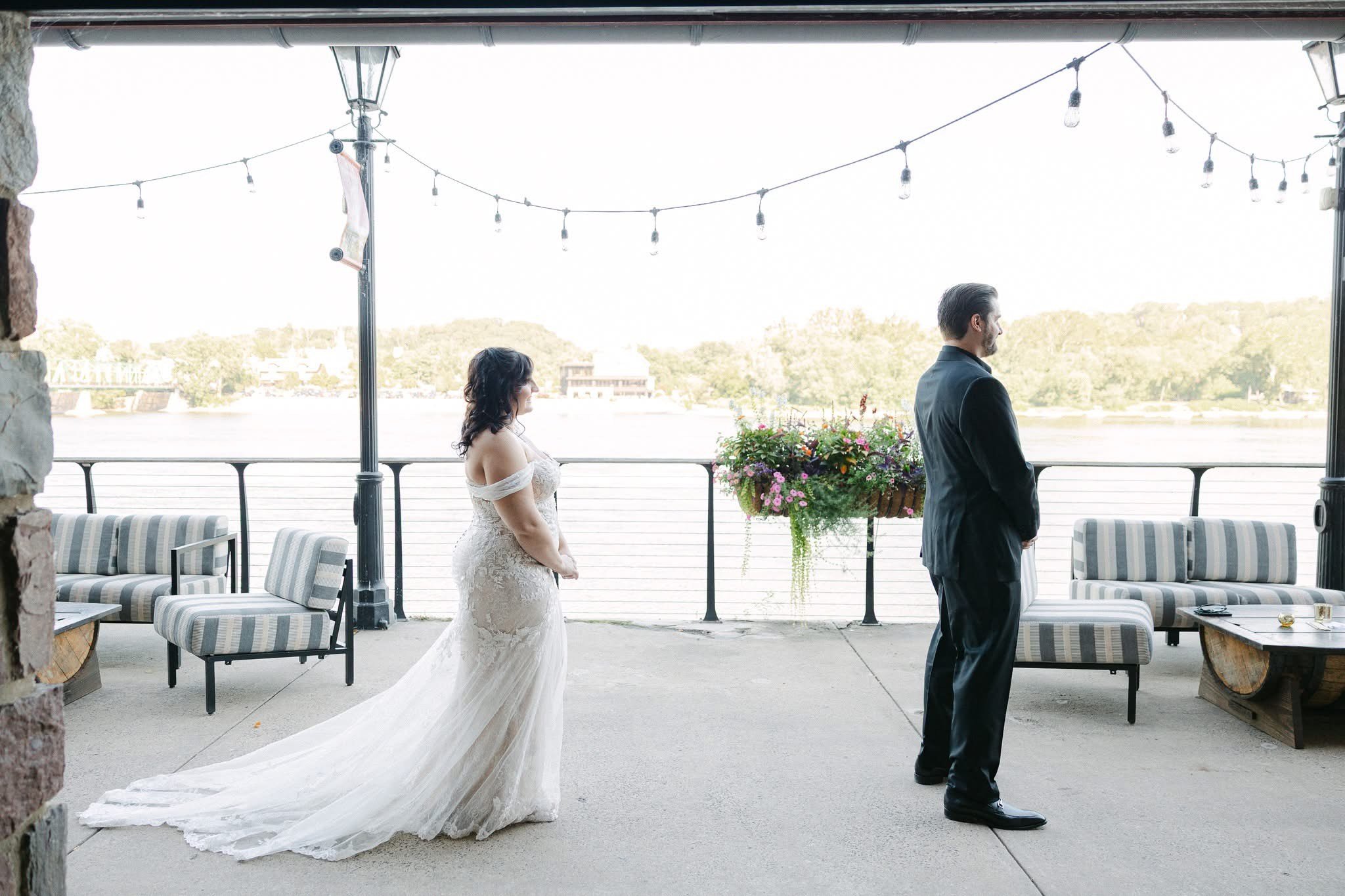 Bride in a white wedding dress standing with her back to groom in a black suit outdoors near a river with string lights overhead and outdoor furniture around.