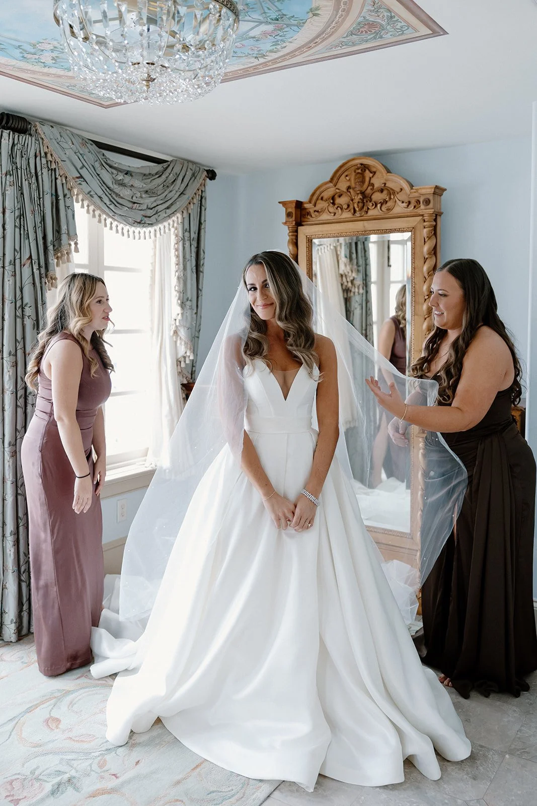 Bride in a white wedding gown and veil standing in a room with two women, one assisting with the veil and another smiling, next to a mirror and window with floral curtains. strapless satin ballgown wona concept bridal dress