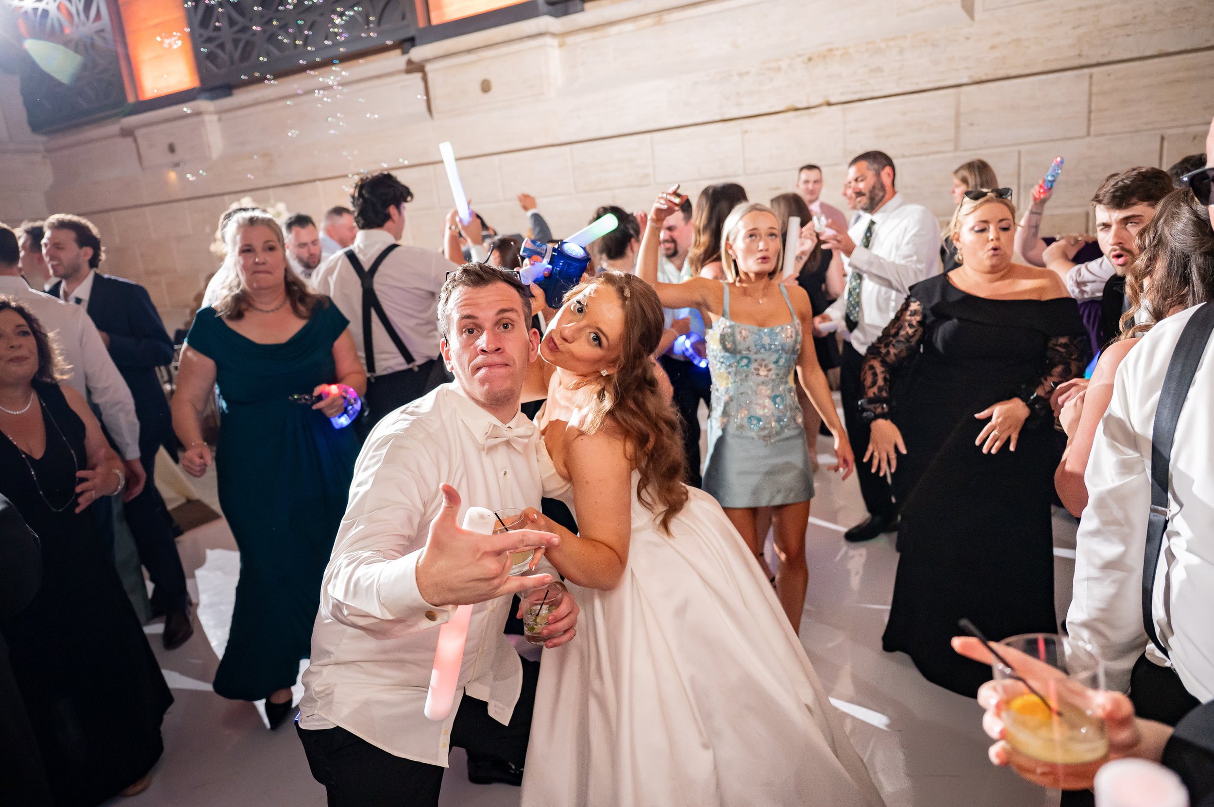 A lively wedding reception with guests dancing, drinking, and celebrating. The bride and groom are in the center, with the groom making a gesture and the bride leaning on him. People are smiling, holding glow sticks, and enjoying the party in a decor