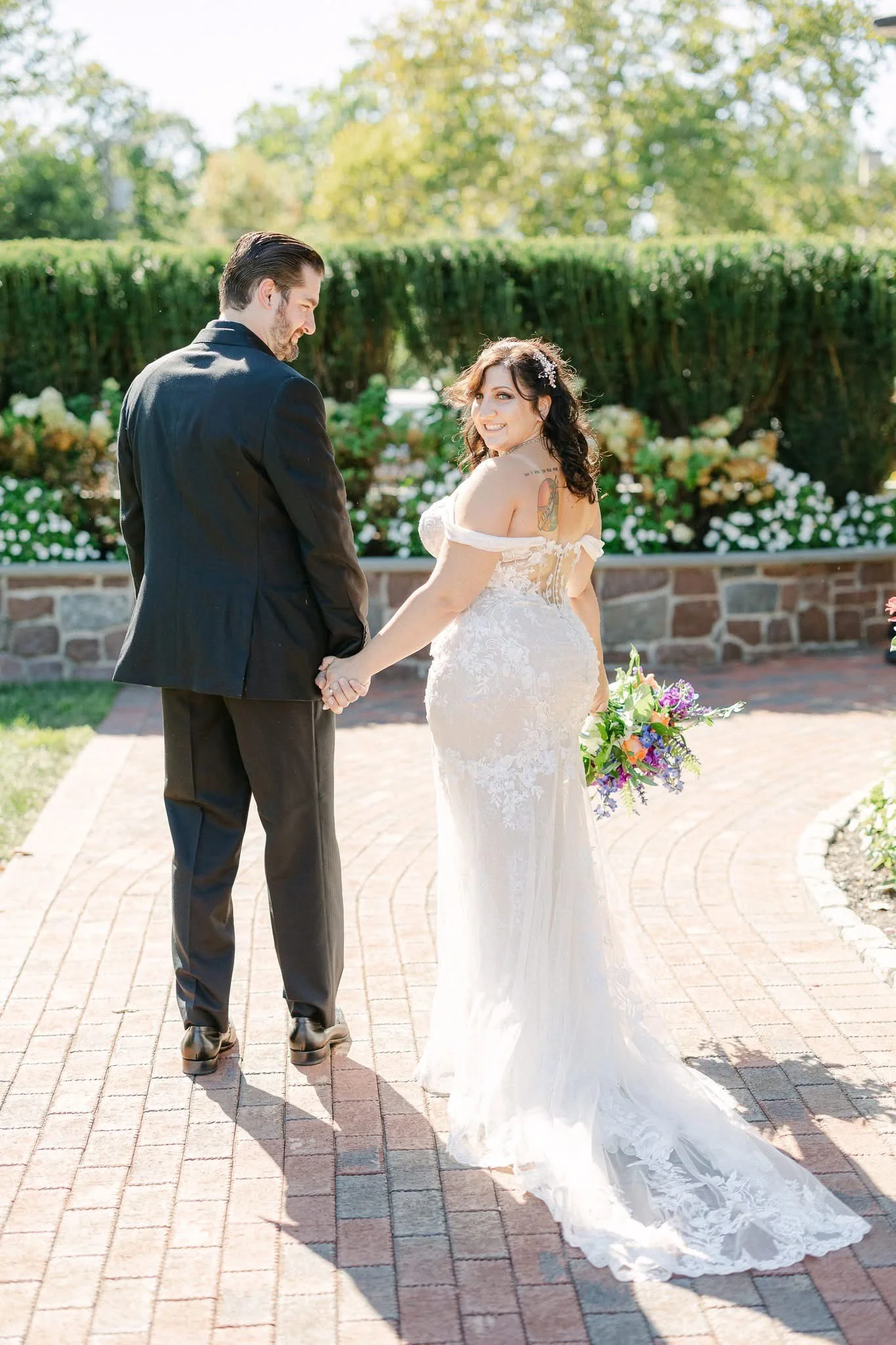 A bride and groom holding hands outdoors on a brick pathway, with the bride in a white lace wedding dress and the groom in a black suit, smiling at each other.