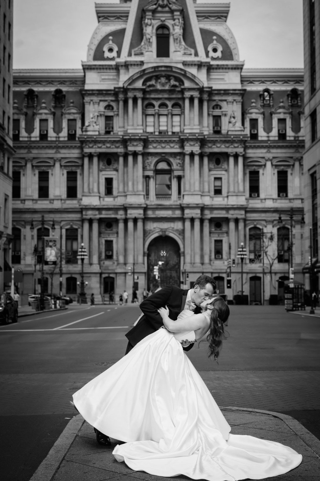 A couple in wedding attire sharing a kiss in front of a grand historic building, Black and white photo.