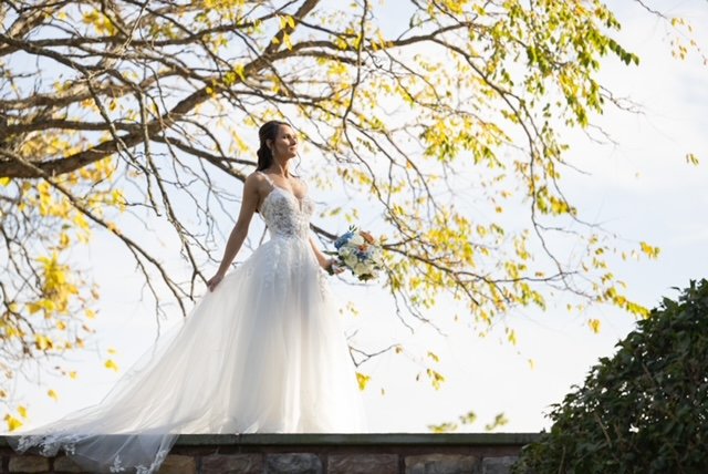 Bride in a white wedding dress holding a bouquet standing outdoors under a tree with yellow leaves.