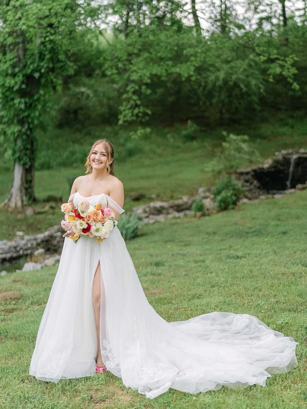 A smiling woman in a wedding dress holding a bouquet of flowers outdoors near a stream, with greenery in the background. Tara Lauren De Mille