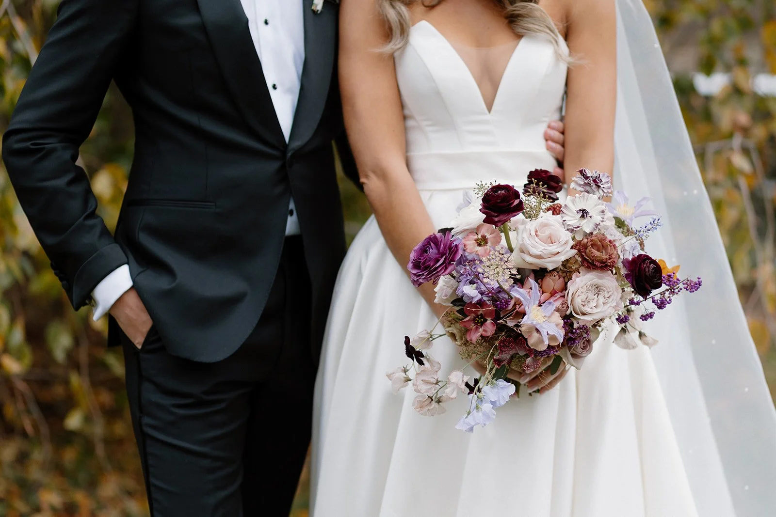 A bride in a white wedding gown holding a colorful bouquet of flowers, standing next to a groom in a black tuxedo, outdoors with autumn foliage in the background. strapless satin ballgown wona concept bridal dress
