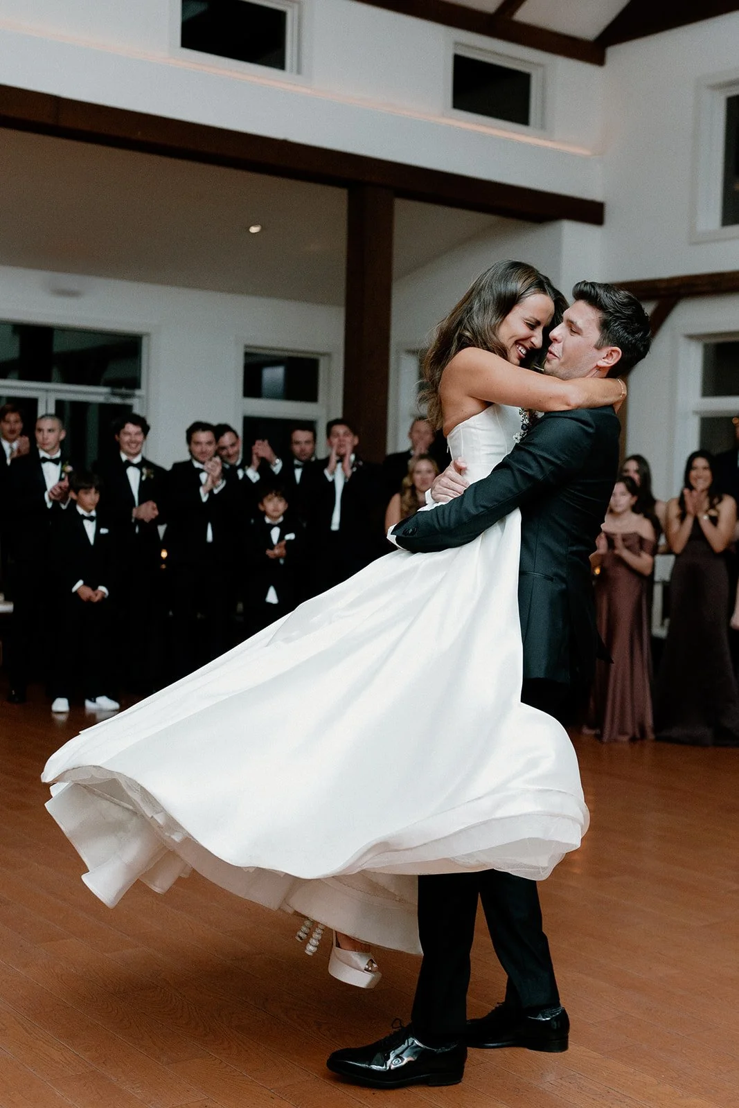 A bride and groom are dancing during their wedding reception inside a spacious room with wooden floors and large windows. The groom lifts the bride, who is wearing a white wedding gown, while they smile and enjoy their moment. strapless satin ballgow
