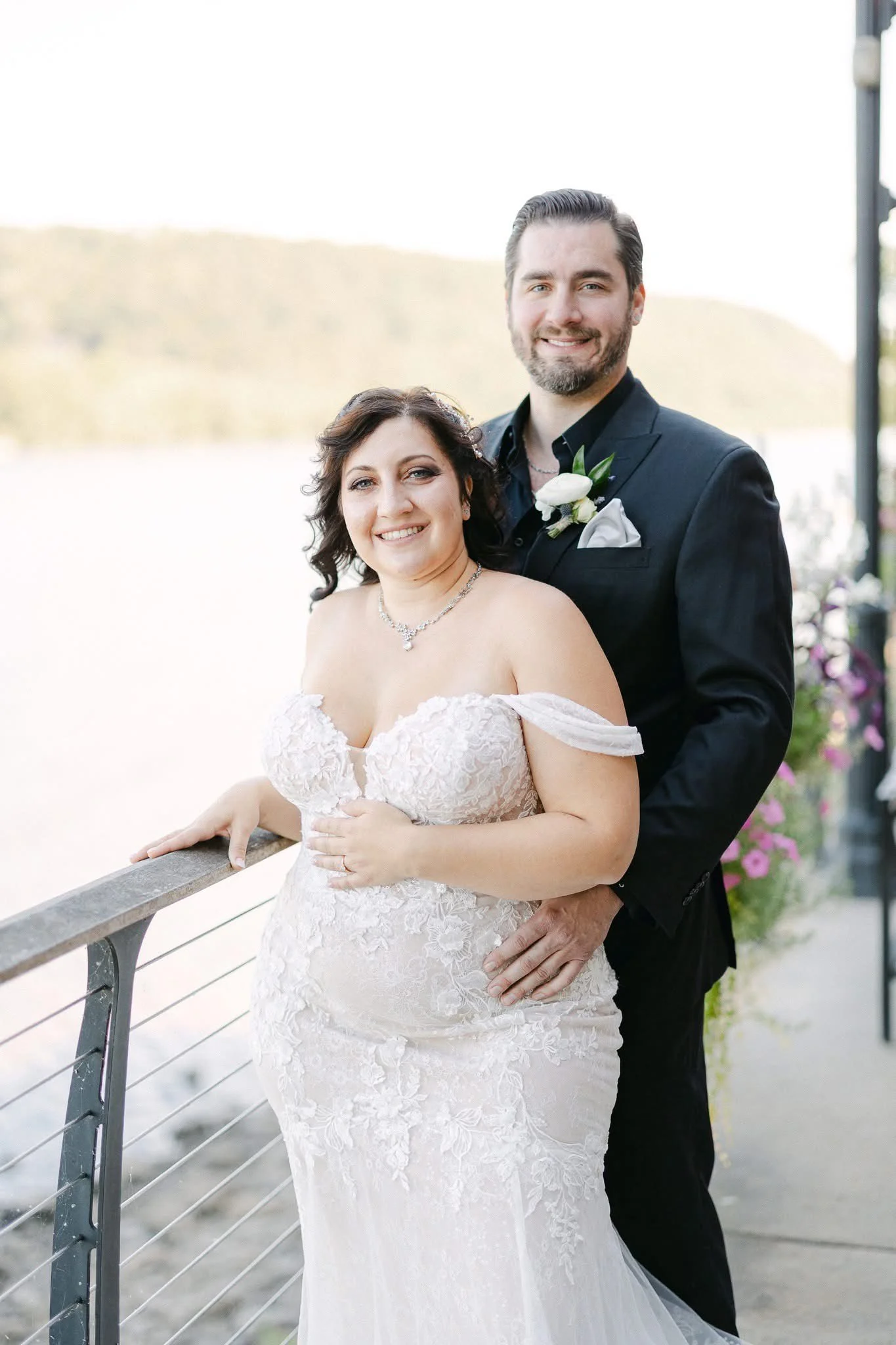 A newlywed couple stands close together outdoors near water, smiling. The bride wears a strapless white lace wedding gown with off-the-shoulder sleeves and a diamond necklace, while the groom wears a black suit with a boutonniere and pocket square.