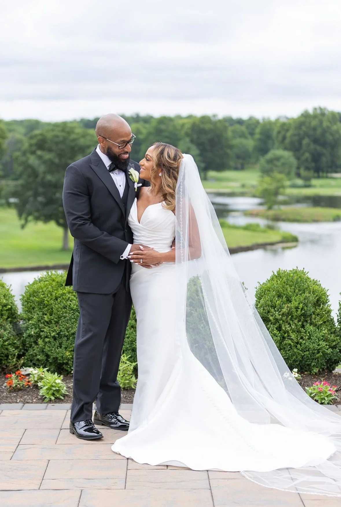 A newlywed couple in wedding attire stands together outdoors, with a scenic lake and green trees in the background. The groom wears a black tuxedo with a bow tie, and the bride wears a white wedding gown with a long veil. Marilyn by Etoile by Elysee
