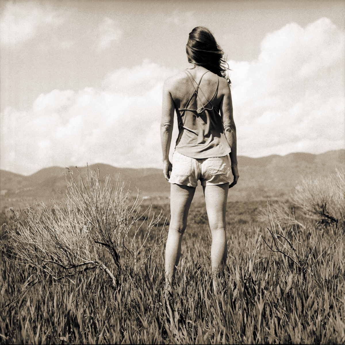 self portrait on a grassy plains, with the mountains in the distance. Photo shows my back and I face the vast open space, hair blowing off to one side,