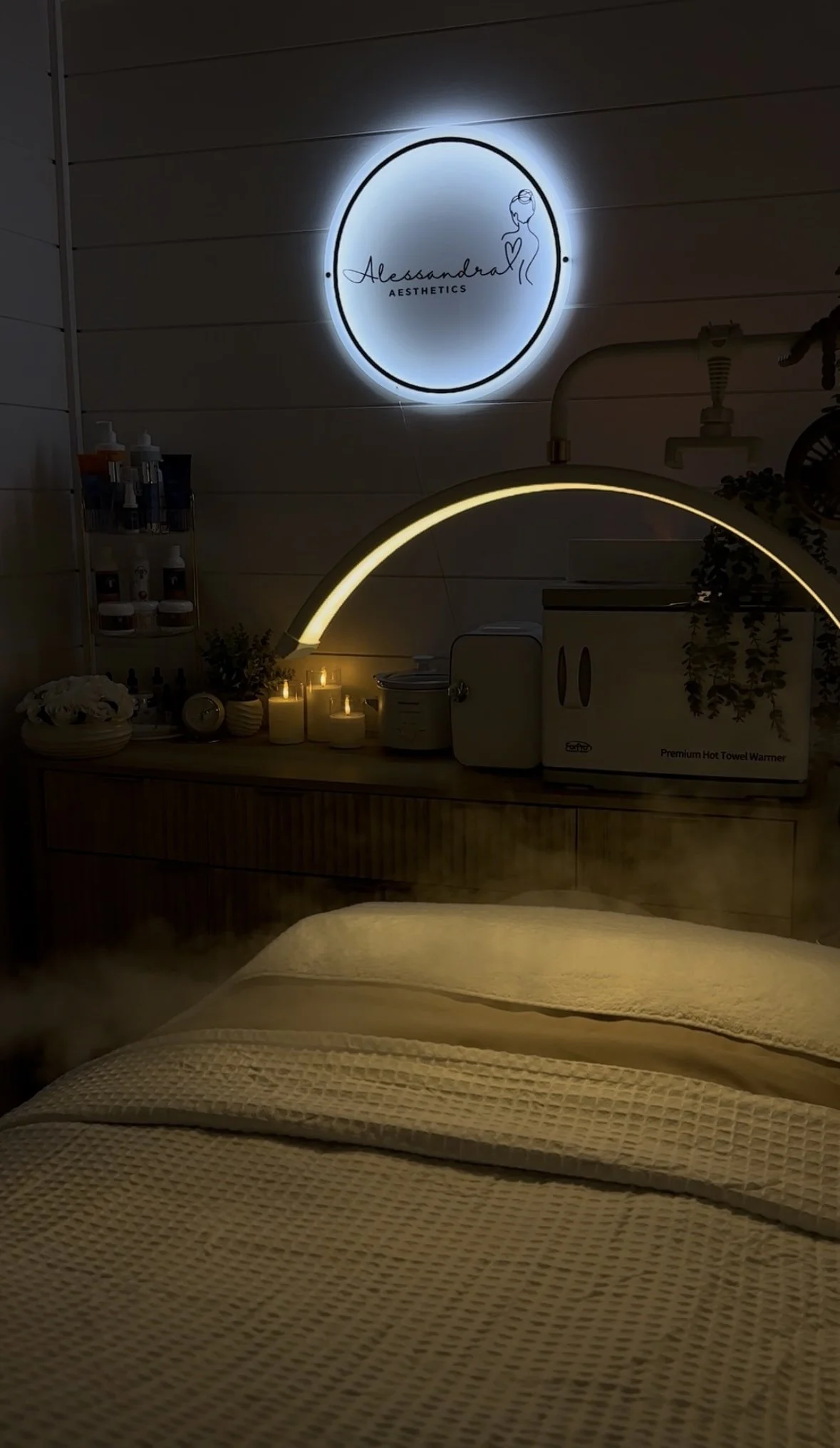 A cozy room in a massage or spa studio with a bed, candles, and skincare products, illuminated with soft lighting, including a neon sign of 'Alessandra Aesthetics' on the wall.