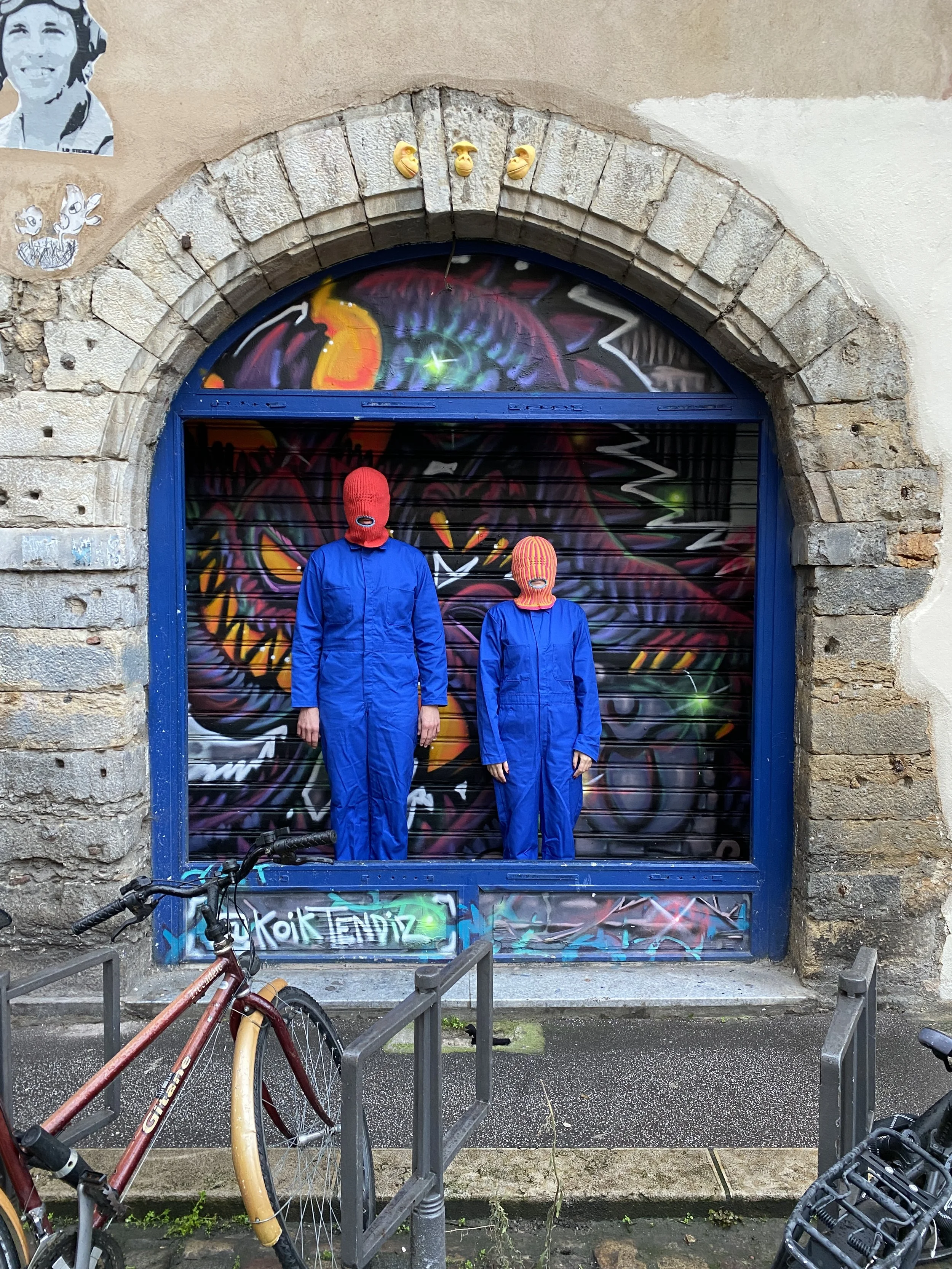 Two Brother Mouth figures in blue outfits and Mouth-Clavas standing in front of a colorful graffiti-covered doorway.