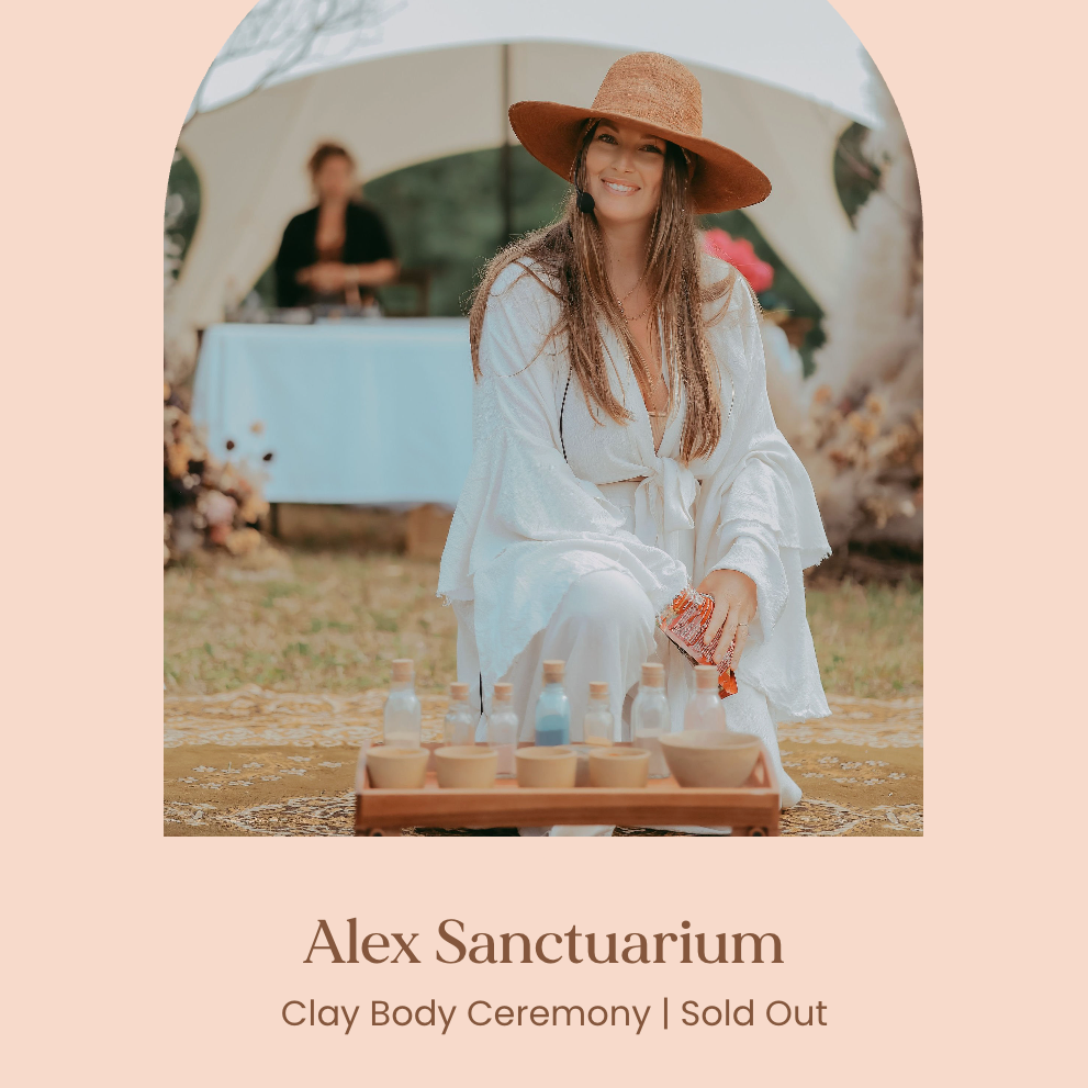 A woman sitting in front of a table with pottery and bottles, outdoors during daytime, wearing a large brown hat and a white dress, smiling. In the background, a person stands behind a table with a white tablecloth at an event, with a tent and floral decorations visible.